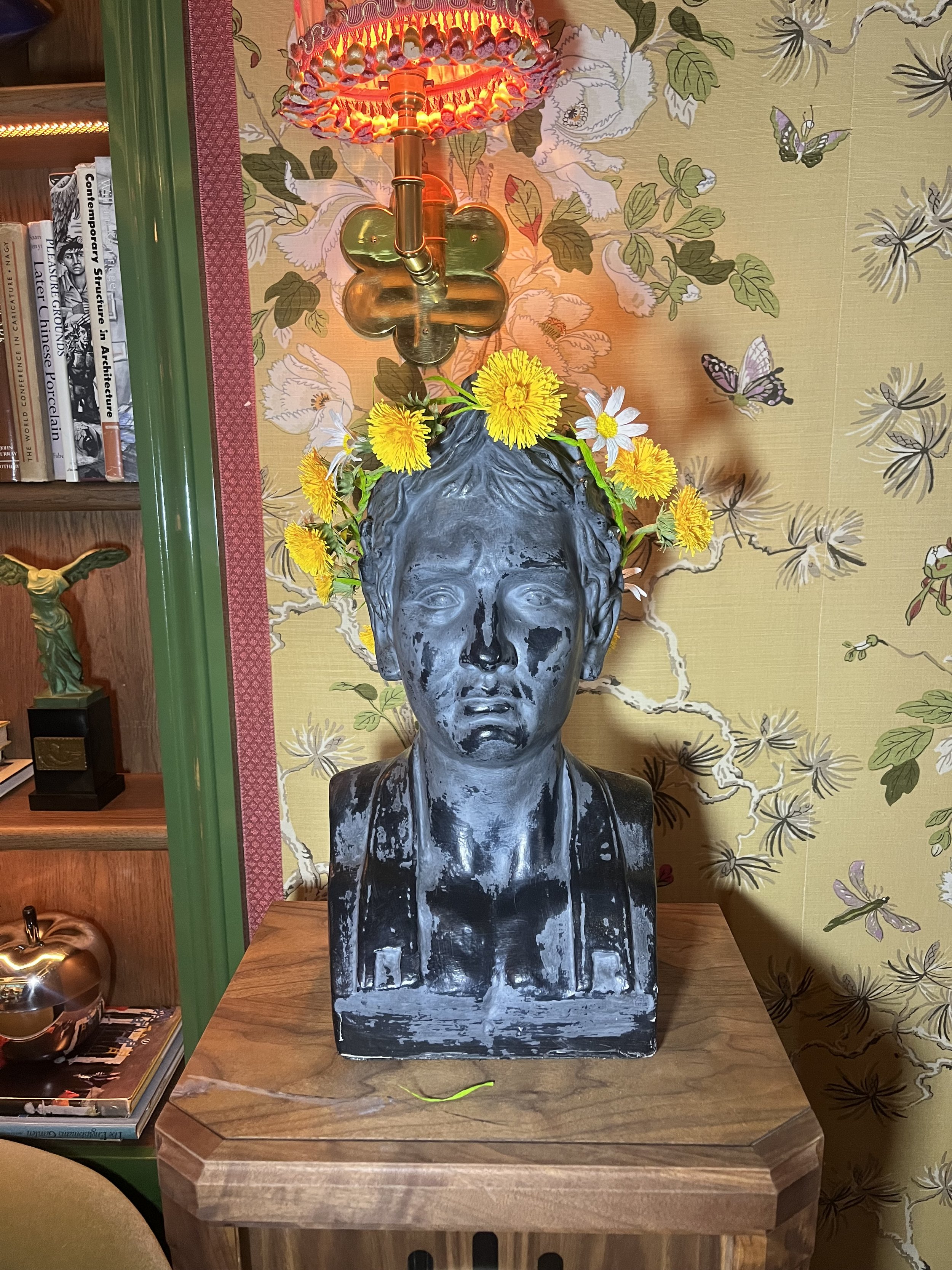 A bust sculpture of a person's head and upper shoulders painted in a distressed black and white finish, placed on a wooden table. The sculpture is topped with a bouquet of yellow and white flowers. Behind the sculpture, a wall features floral and but