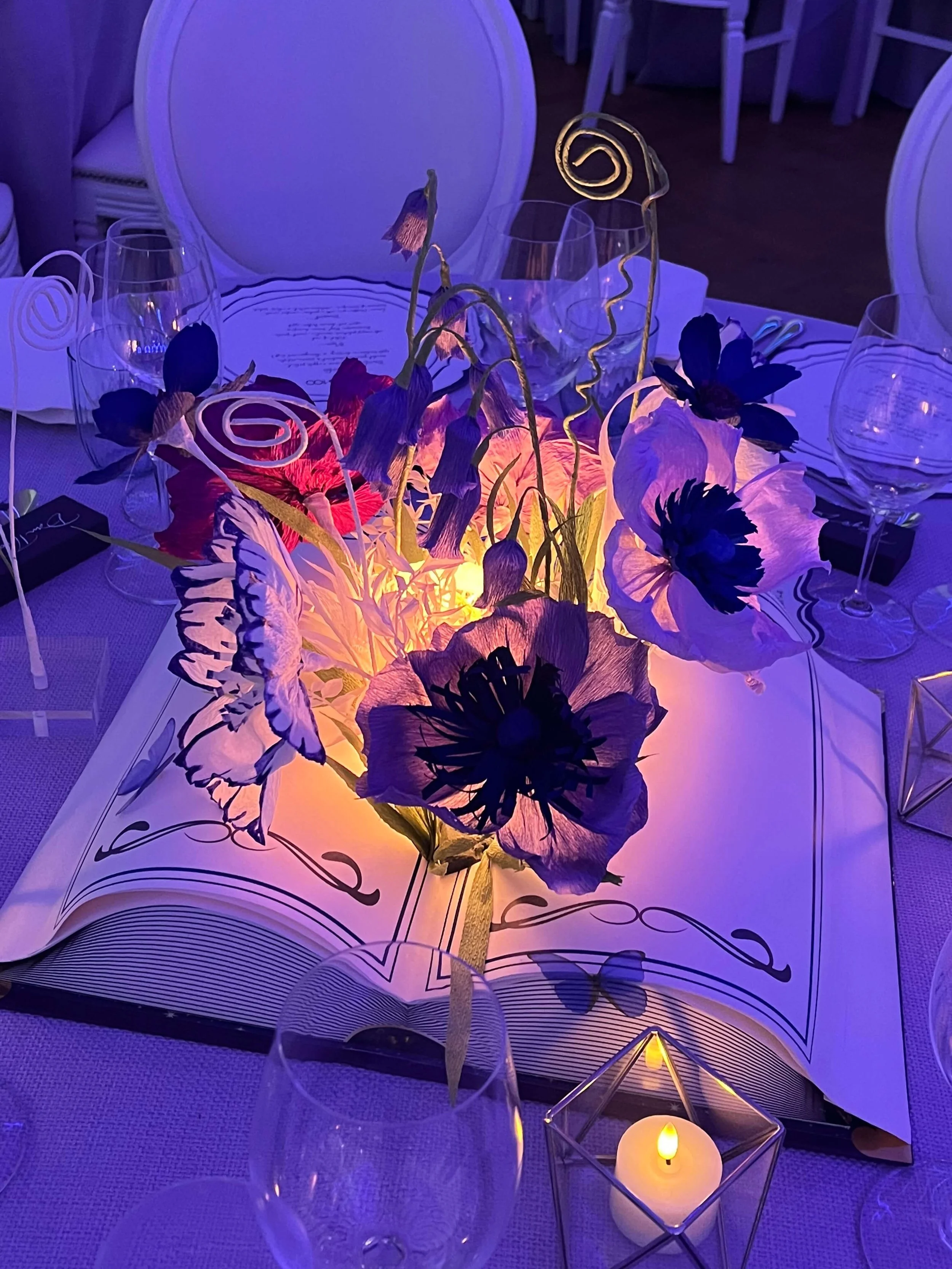 A dinner table decorated with an arrangement of purple and pink paper flowers by Emmeli Kimhi for Disney, illuminated by warm lighting and candle flames inside a glass holder, with empty wine glasses and a menu on the table.