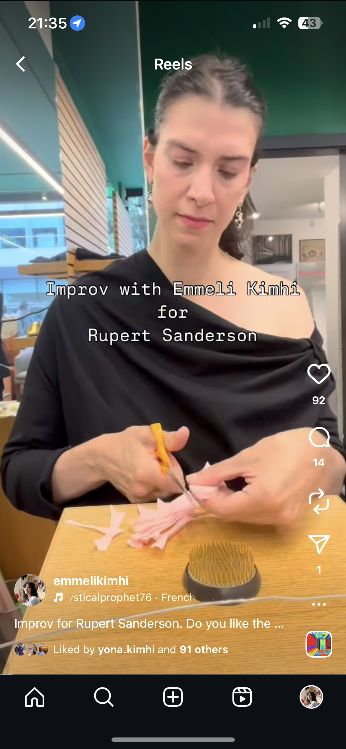 Emmeli Kimhi using scissors to cut pink fabric. There is a yellow-handled tool in her left hand and a small brush with stiff bristles on the table. The background shows a modern interior with wooden accents and large windows.