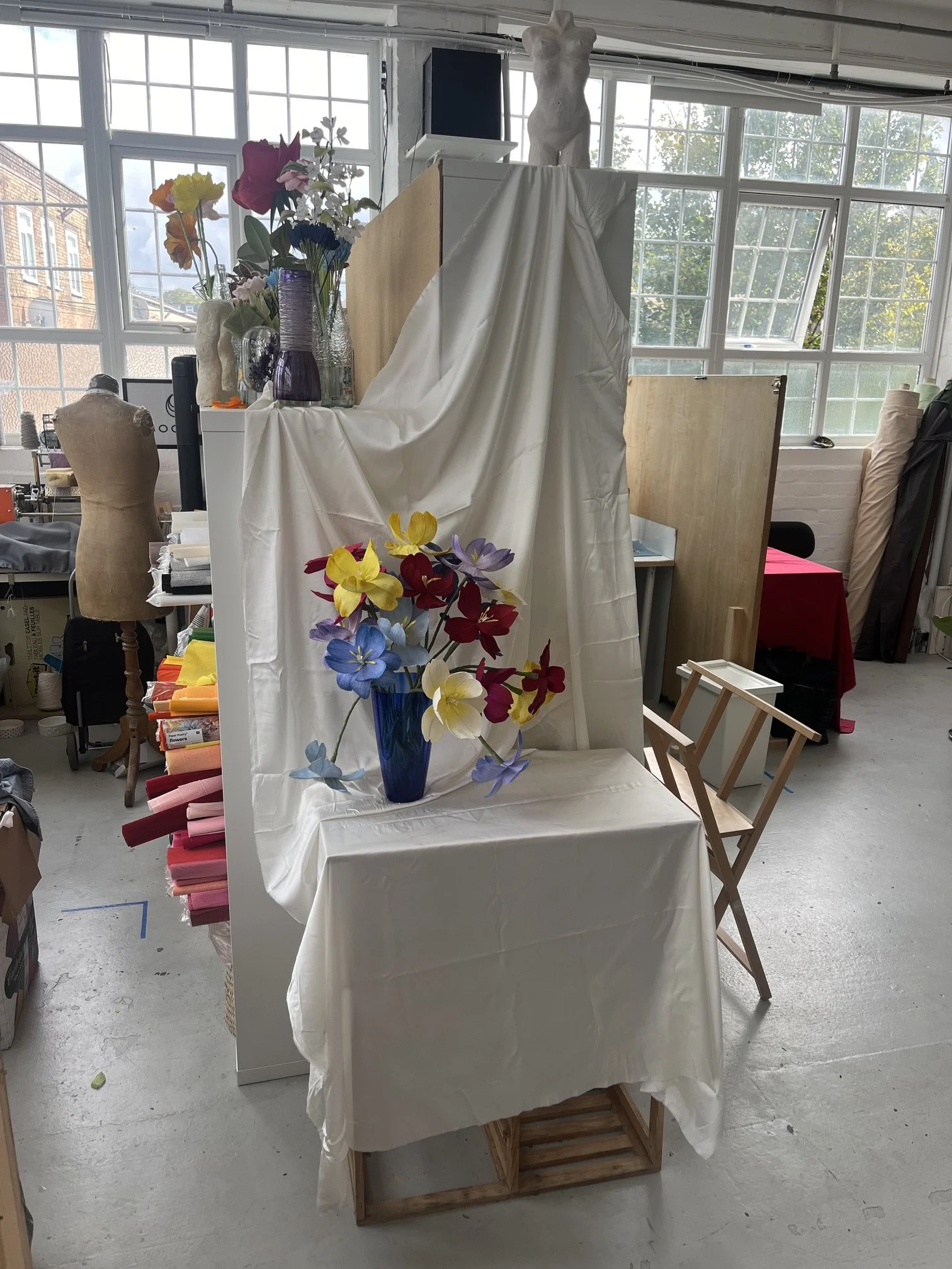 Emmeli Kimhi's art studio with large windows, displaying a mannequin, a flower arrangement in a blue vase, and a draped white sheet with more flowers. Various art supplies and tools are scattered across the workspace.