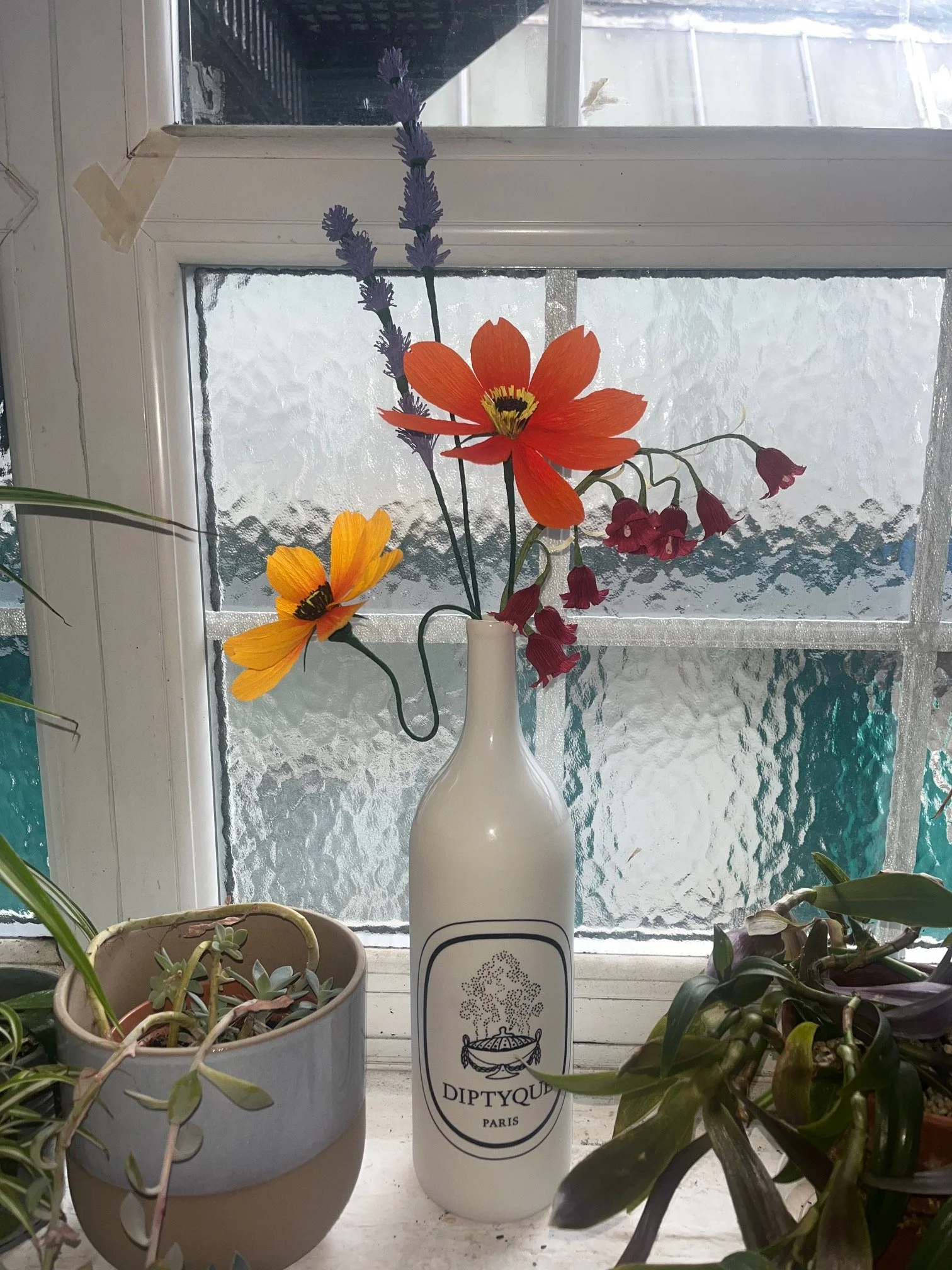 A white vase with the Diptyque Paris logo holds a bouquet of purple lavender, a yellow and orange flower, and red bells by Emmeli Kimhi, placed on a windowsill surrounded by various potted plants, with frosted glass window in the background.