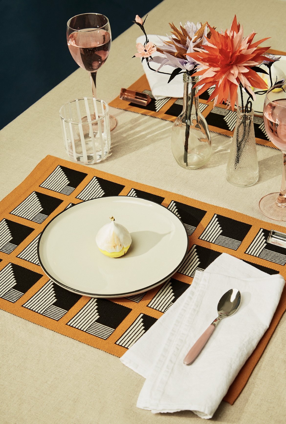 A table setting with a beige tablecloth, decorated with orange and black geometric placemats, a white plate with a small dessert, a silver spoon on a white napkin, two glasses of pink wine, and three glass vases with pink and orange paper flowers.
