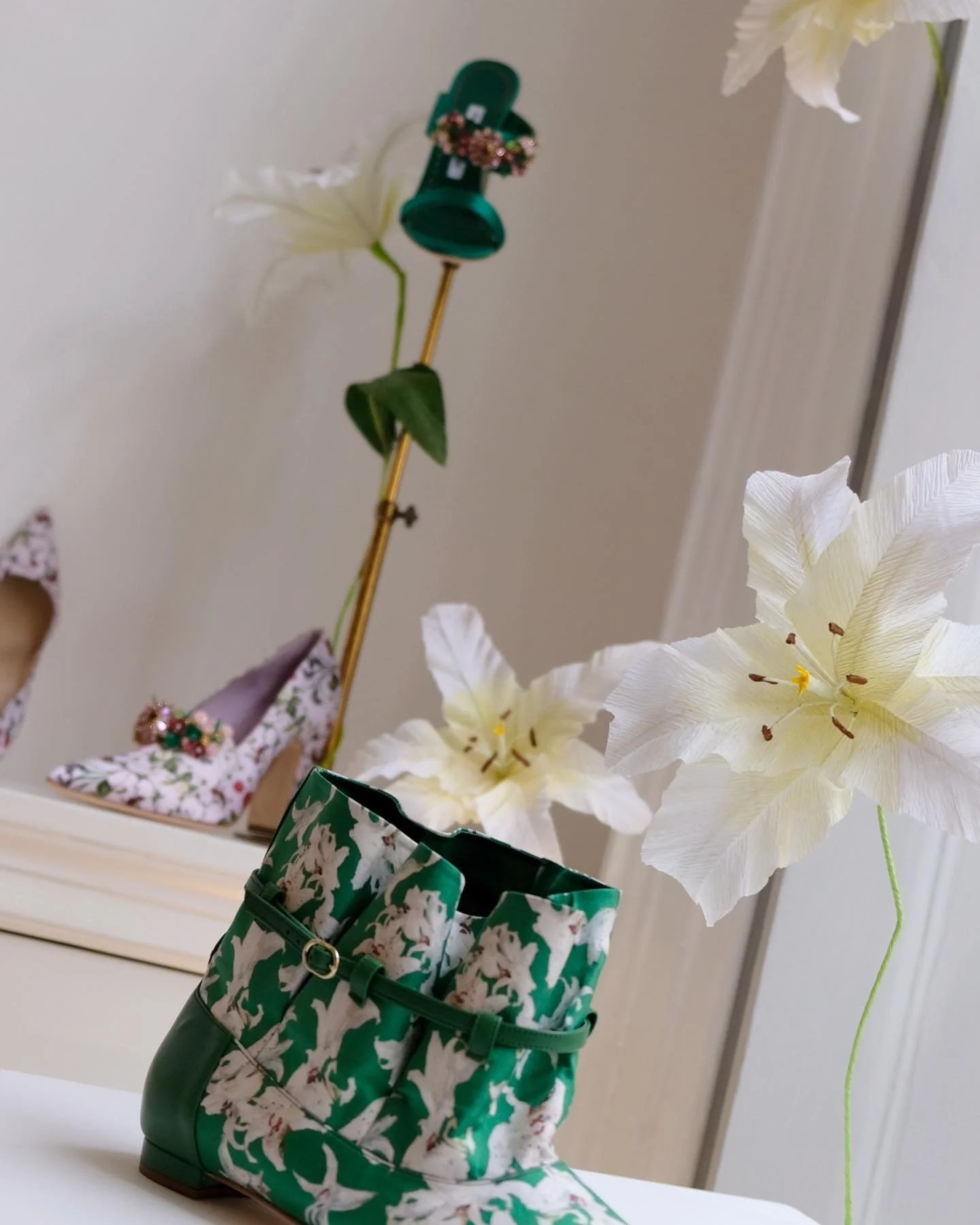A decorative display featuring white paper lilies by Emmeli Kimhi in a green and white patterned rain boot, with a floral patterned high-heeled shoe in the background.