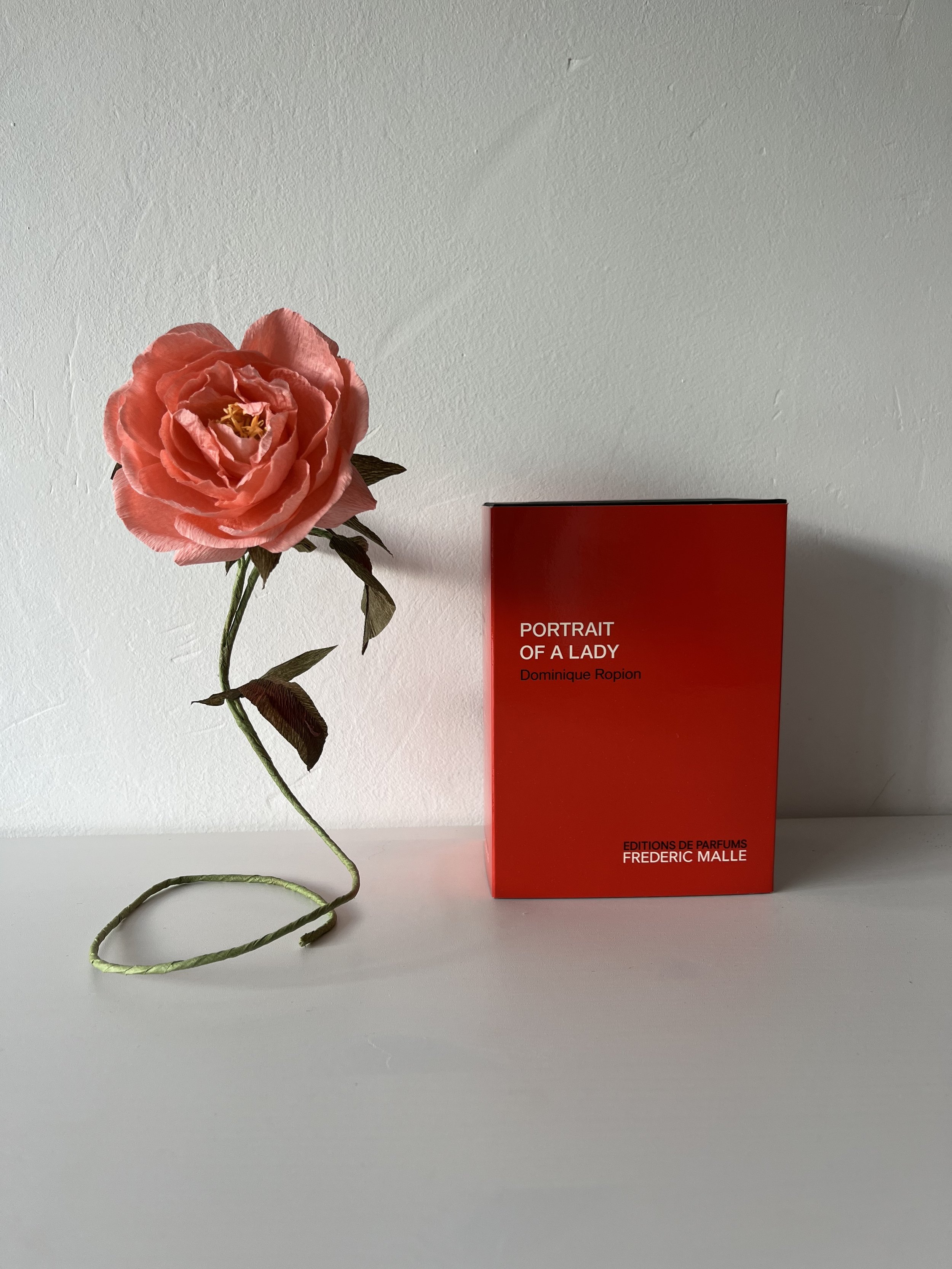 A pink artificial rose  by Emmeli Kimhi with dark green leaves and stem placed next to a red book titled 'PORTRAIT OF A LADY' by Frederic Malle , set against a plain white background.