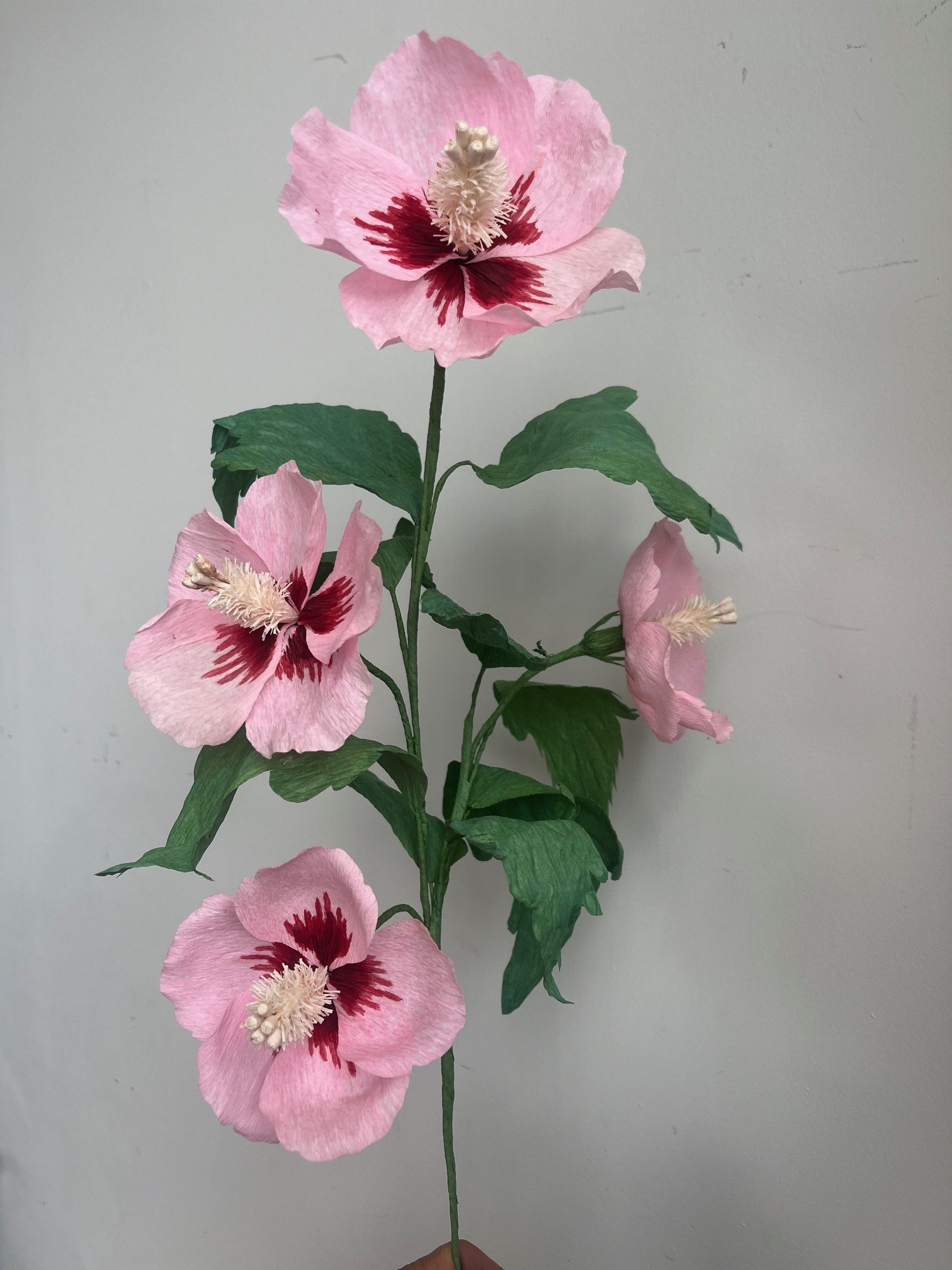 Artificial pink hibiscus flower by Emmeli Kimhi with dark red markings and white stamens, with green leaves on a plain light gray background.