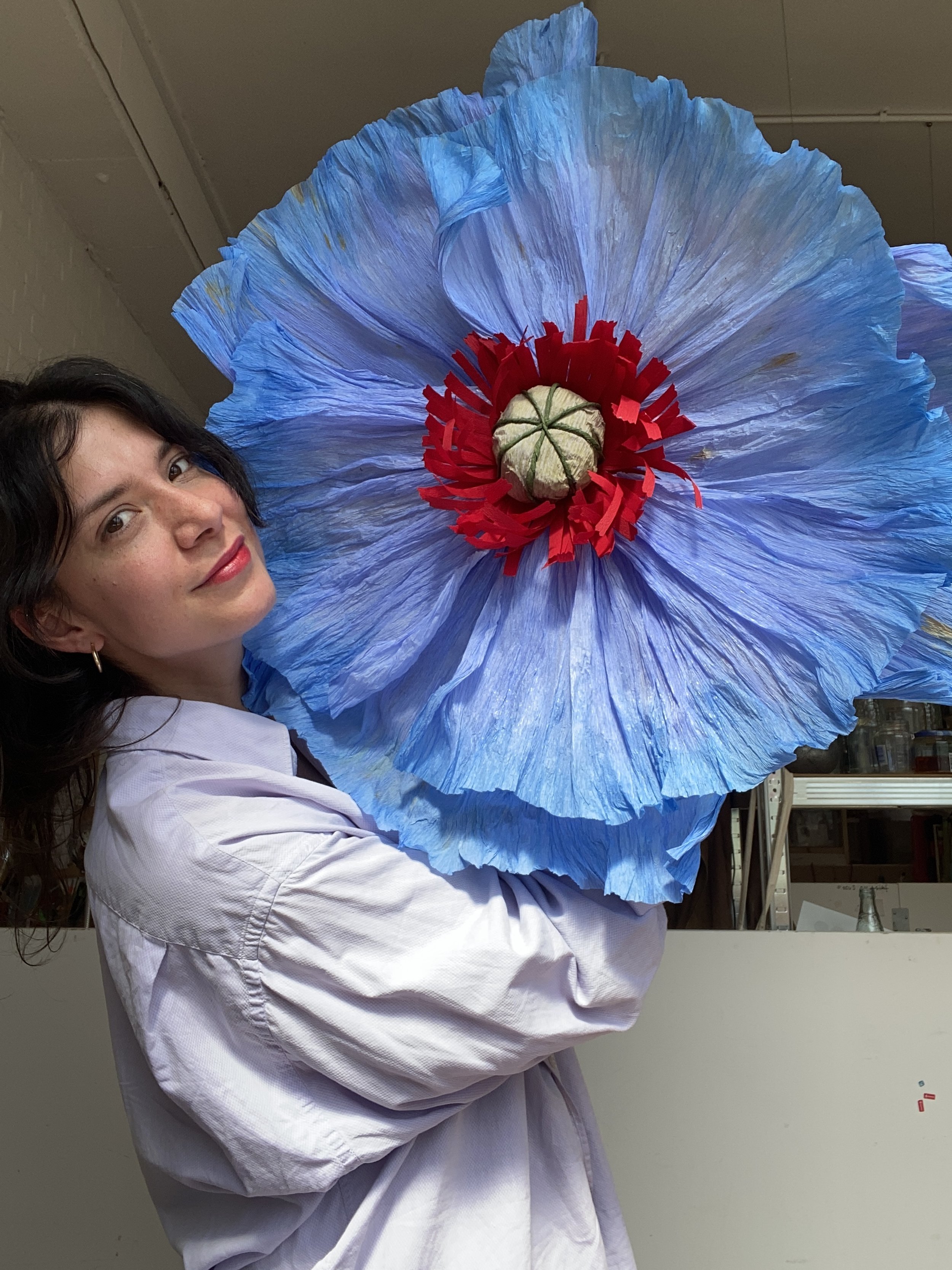 Emmeli Kimhi with dark hair, wearing a white shirt, holding a large blue paper flower with a red and white center.