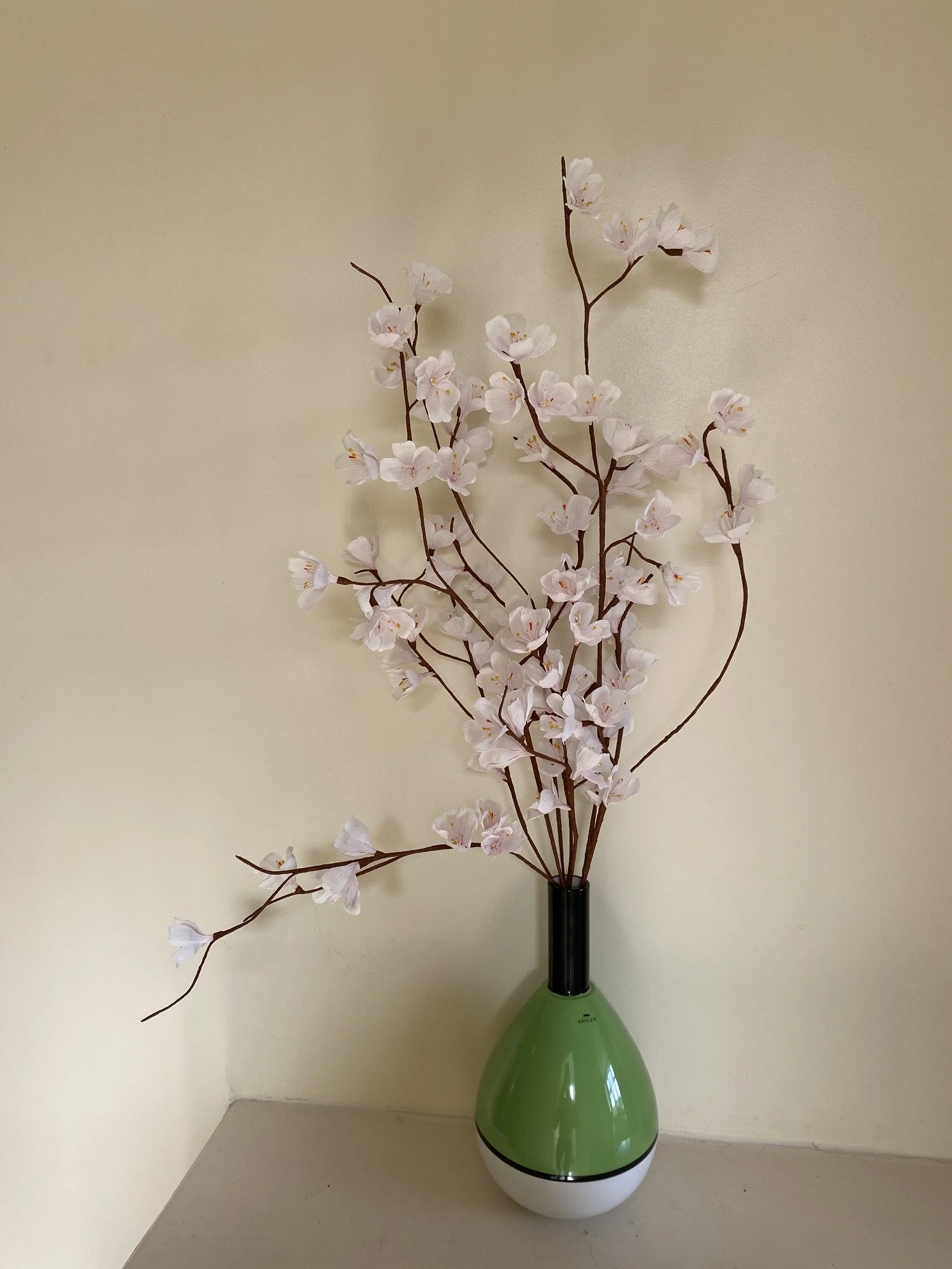 Artificial cherry blossom flowers by Emmeli Kimhi in a green and white vase placed on a beige table against a plain beige wall.