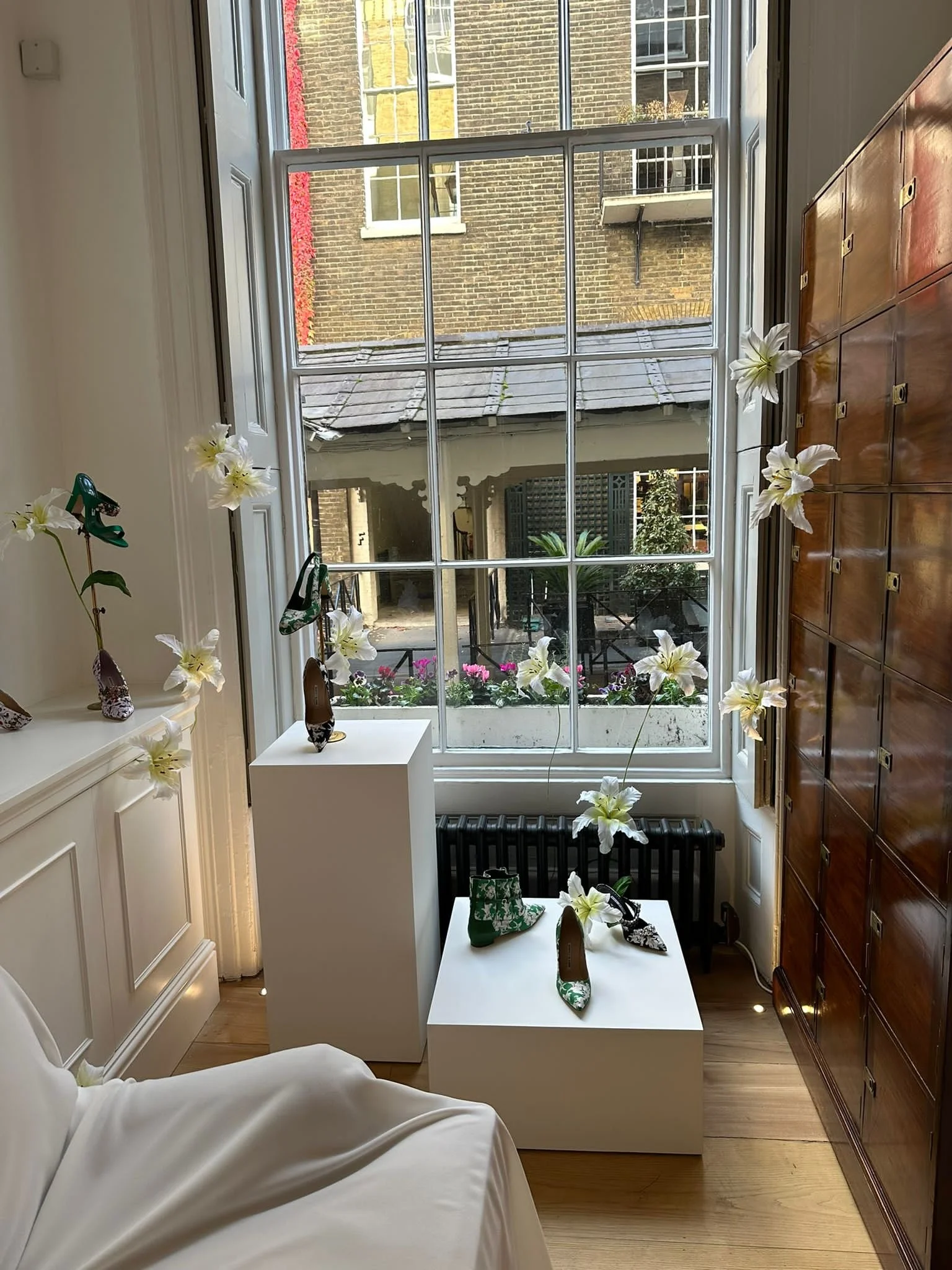 Interior of a room with a large window overlooking a city exterior, decorated with white paper lilies  by Emmeli Kimhi and display shoes on white pedestals, and a wooden locker on the right side.