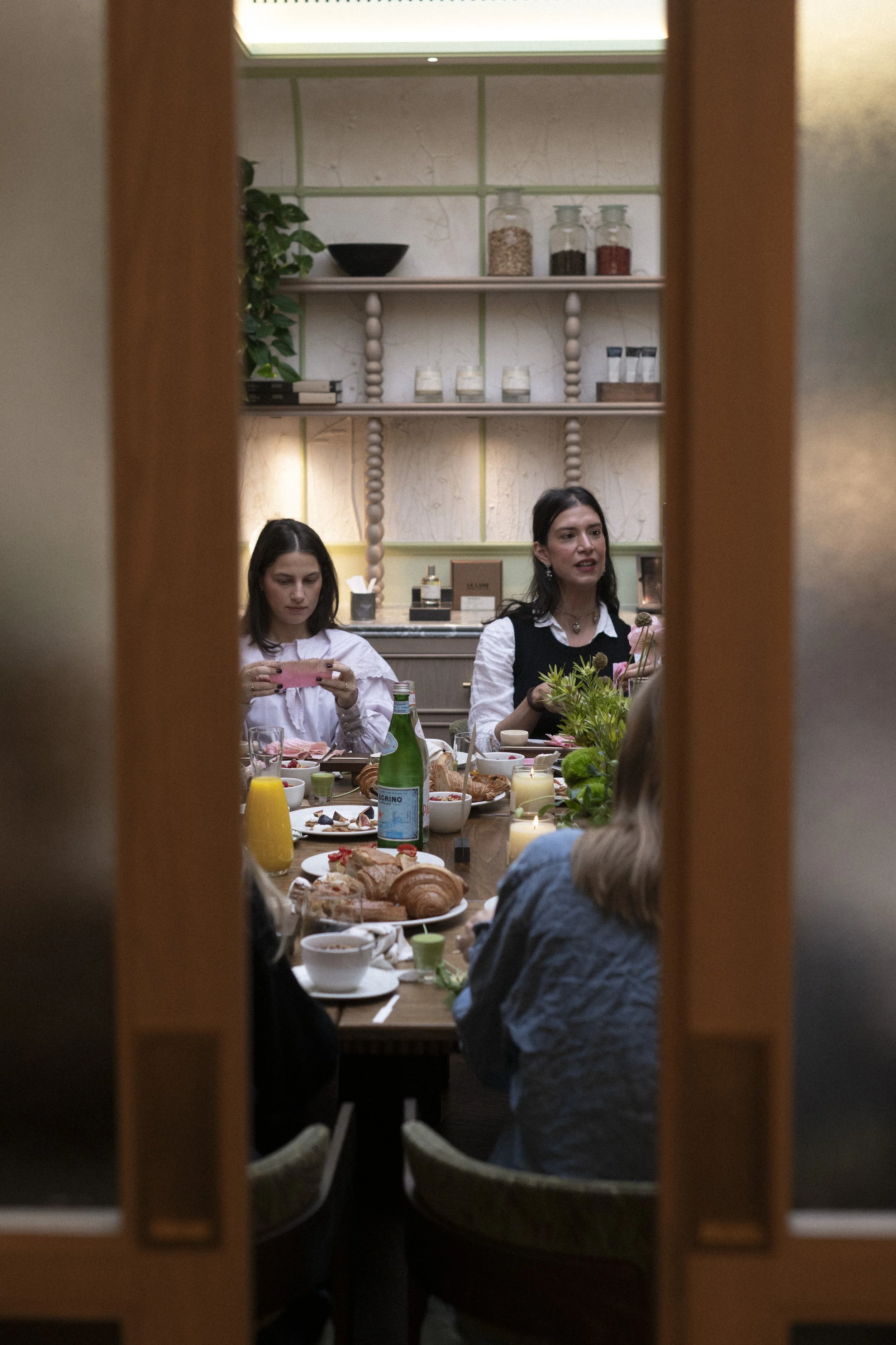 Emmeli Kimhi with a group of people having a meal at a dining table, seen through a door frame, with food and drinks on the table, in a cozy, decorated room. Workshop.