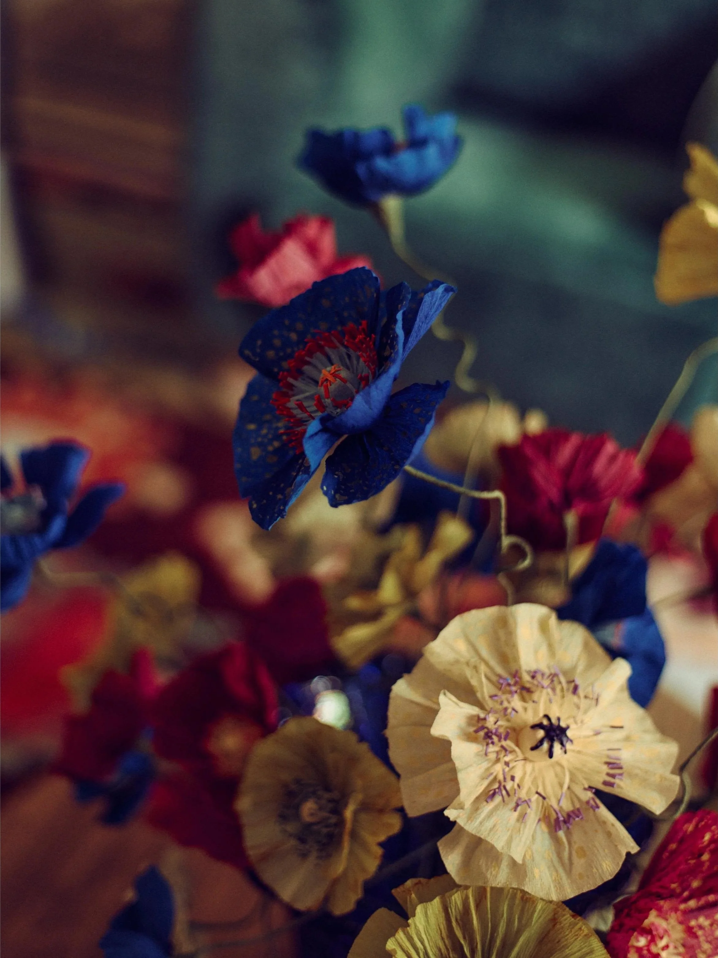 Close-up of colorful paper flowers by Emmeli Kimhi, primarily blue, yellow, and red, with ruffled petals and detailed centers, arranged together. At Alcova Milan.