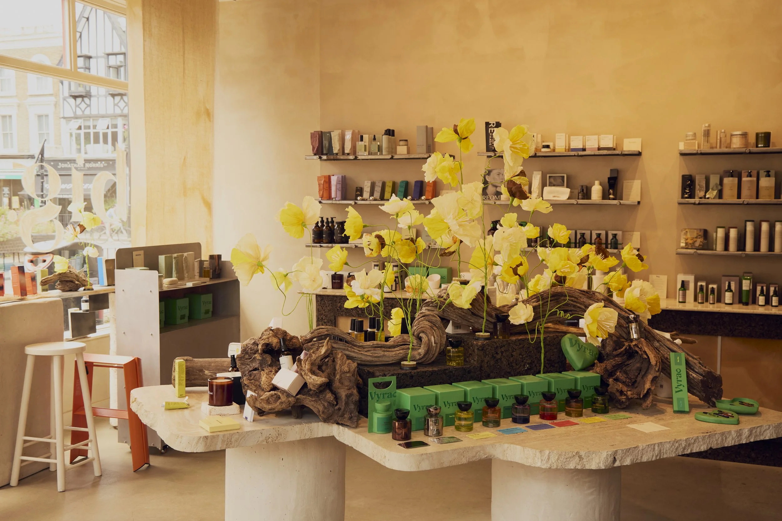 Interior view of a boutique store with shelves of beauty and skincare products, a central display with decorative yellow flowers and natural wood accents, large window on the left letting in natural light.