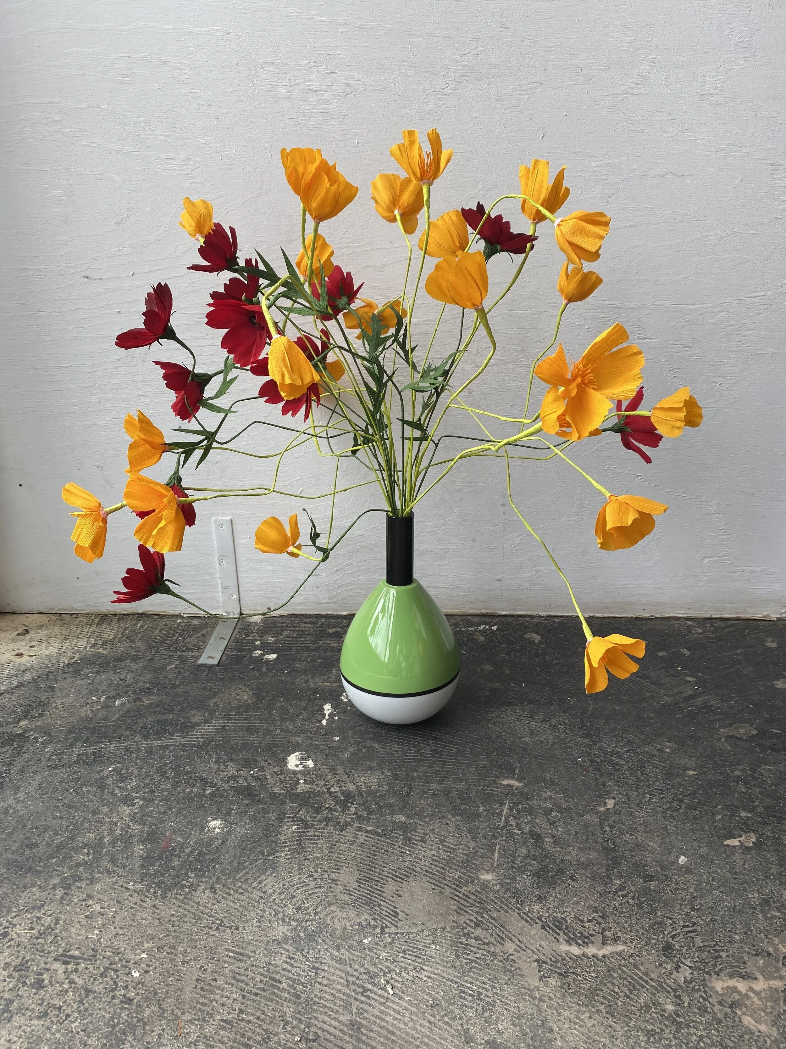A bouquet of red and orange paper flowers flowers by Emmeli Kimhi in a green and white vase placed on a dark, textured floor against a plain neutral wall background.