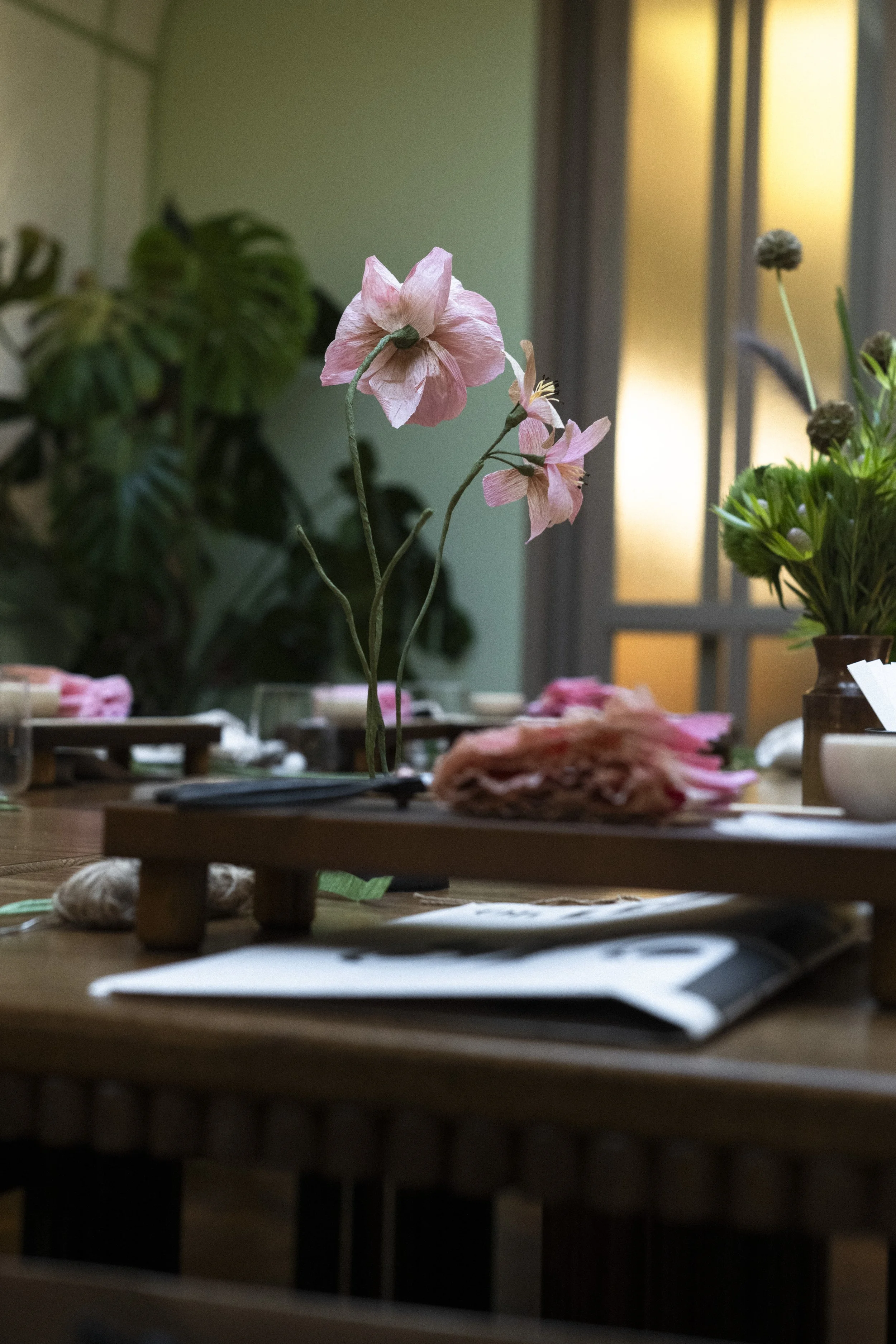Emmeli Kimhi workshop. A pink paper flower centerpiece on a wooden table, with other flowers and tableware in the background.