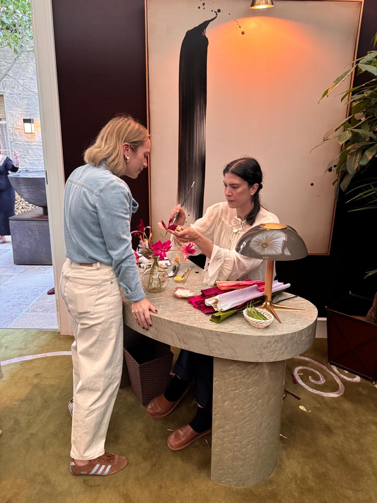 Paper flower artist Emmeli Kimhi demonstrates the creation of paper flowersto Tiffany Duggan at the Black Editions SPeakEasy Salon by Studio Duggan during London's WOW!house designer showcase, surrounded by colourful hand painted paper flowers.