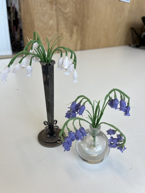 Artificial flowers by Emmeli Kimhi depicting wilted white and purple bell-shaped flowers in a metal and glass vase, placed on a white surface.