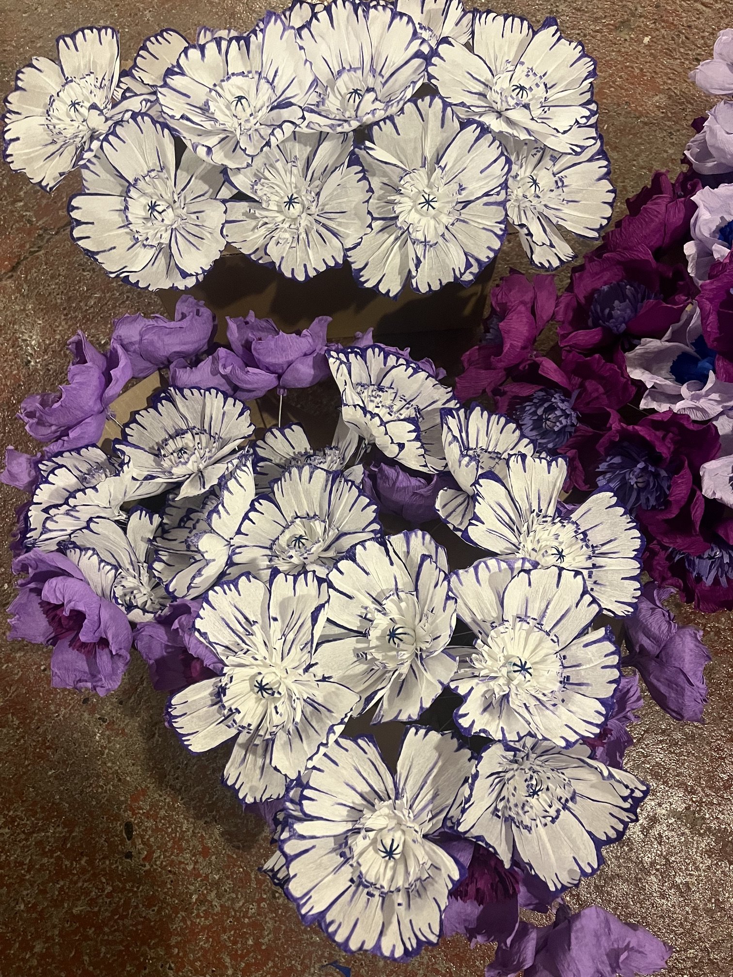 Collection of large artificial flowers by Emmeli Kimhi with white petals outlined in dark purple and purple leaves on a textured surface.