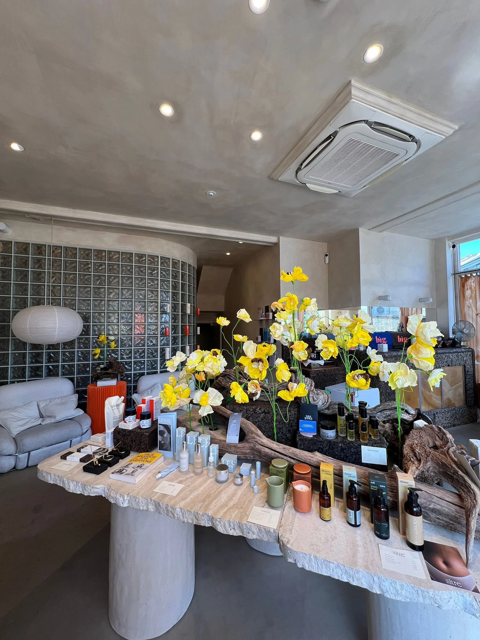 A display table with skincare and beauty products, decorative yellow flowers by Emmeli Kimhi, and informational cards in a boutique or spa setting.