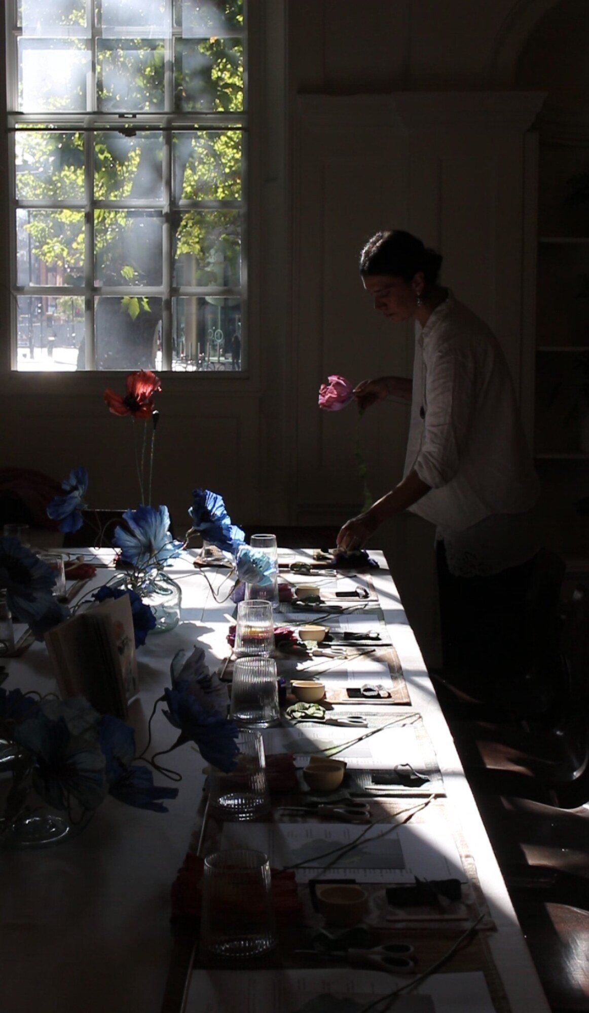 Long workshop table in window light at Blewcoat, set with blue paper poppies in glass vases