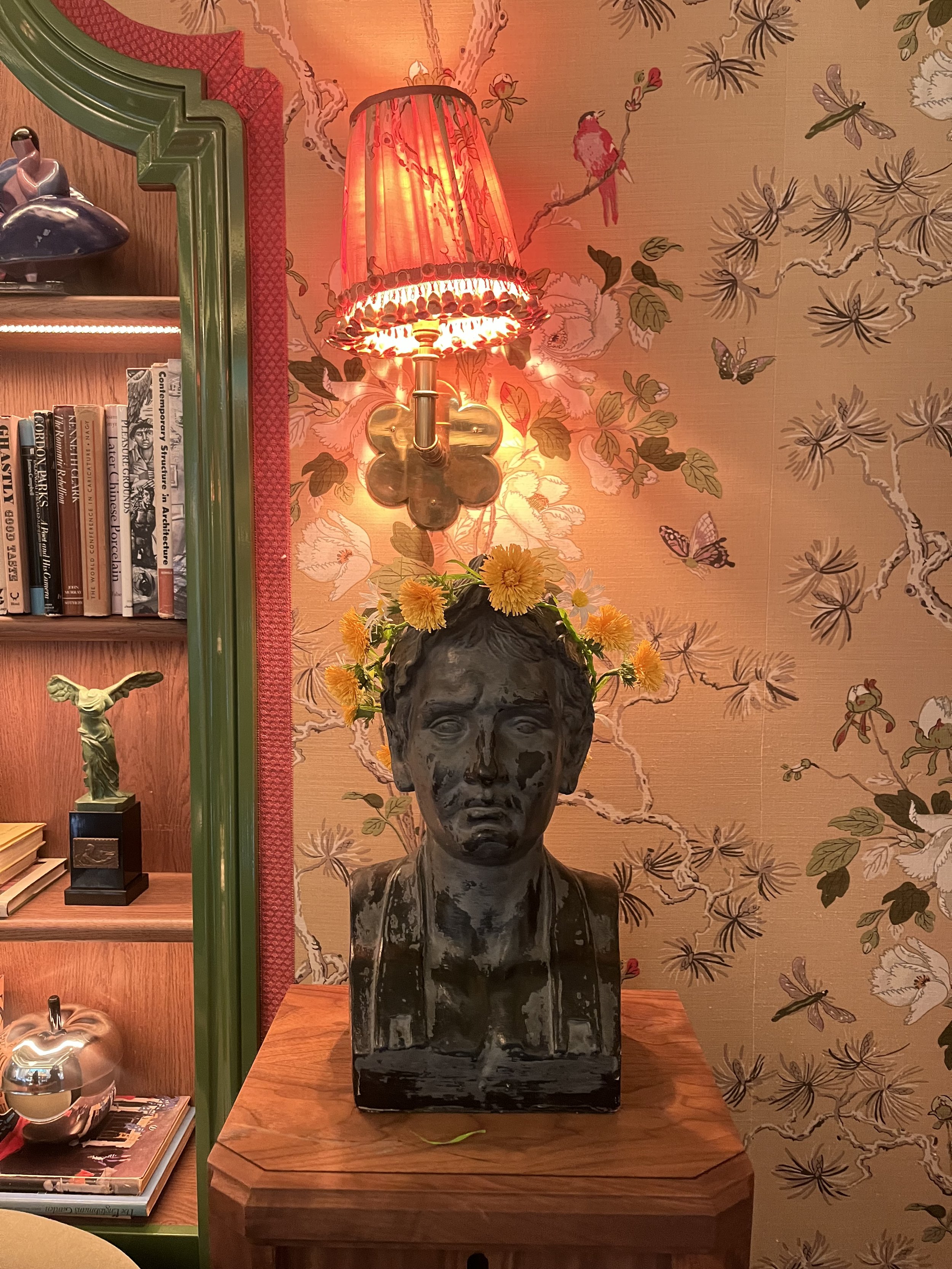 A black bust sculpture of a young boy with flowers on top, placed on a wooden surface. Behind it is a wall decorated with floral and butterfly wallpaper. To the left, there's a bookshelf with books and a small bronze sculpture of an angel. Below the 