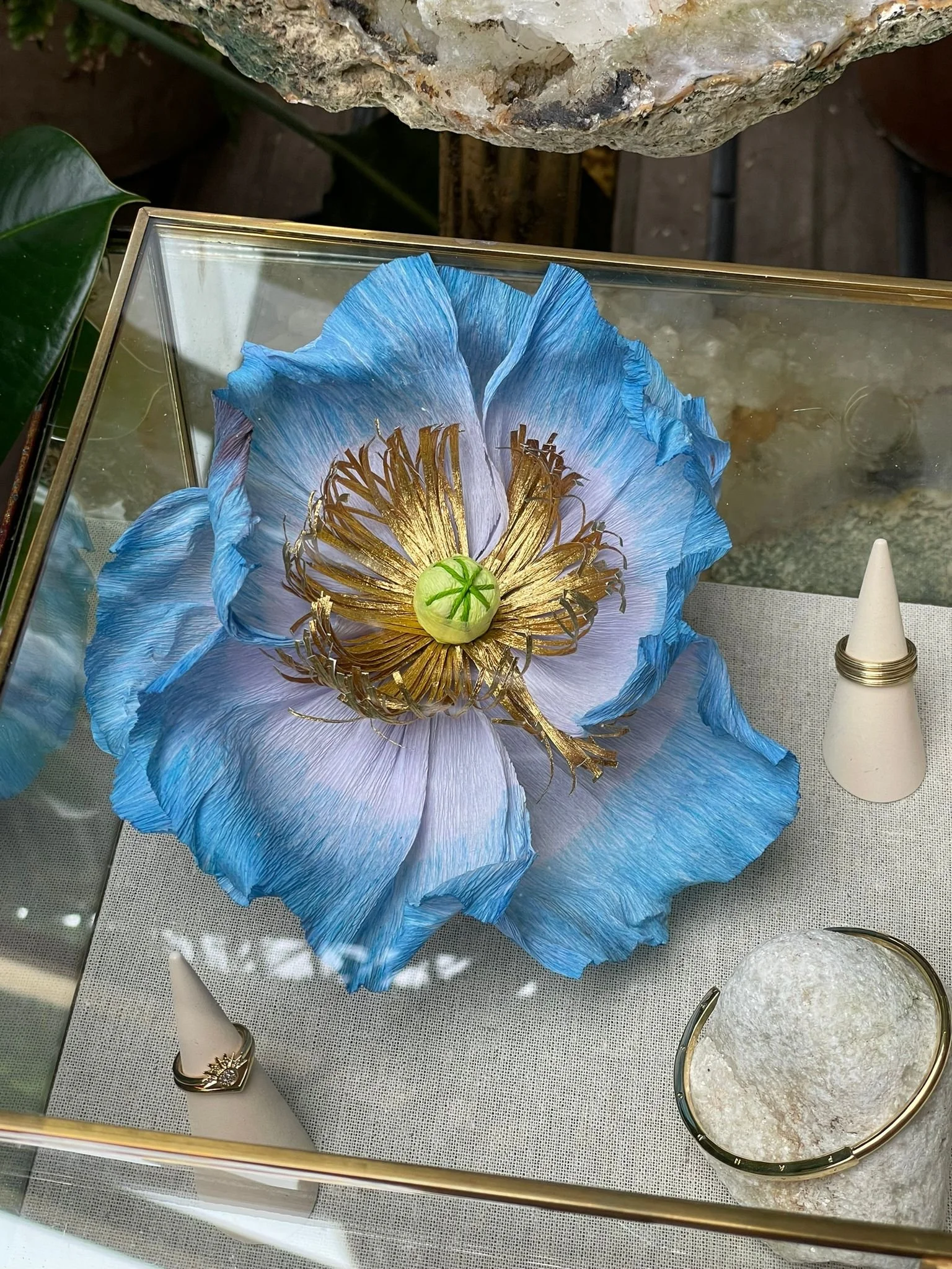 Pandora Display case showing a large blue and gold decorative flower made of paper by Emmeli Kimhi with a green seed-like center. Surrounding items include a ring with a sunburst design and other jewelry, with a rough stone.