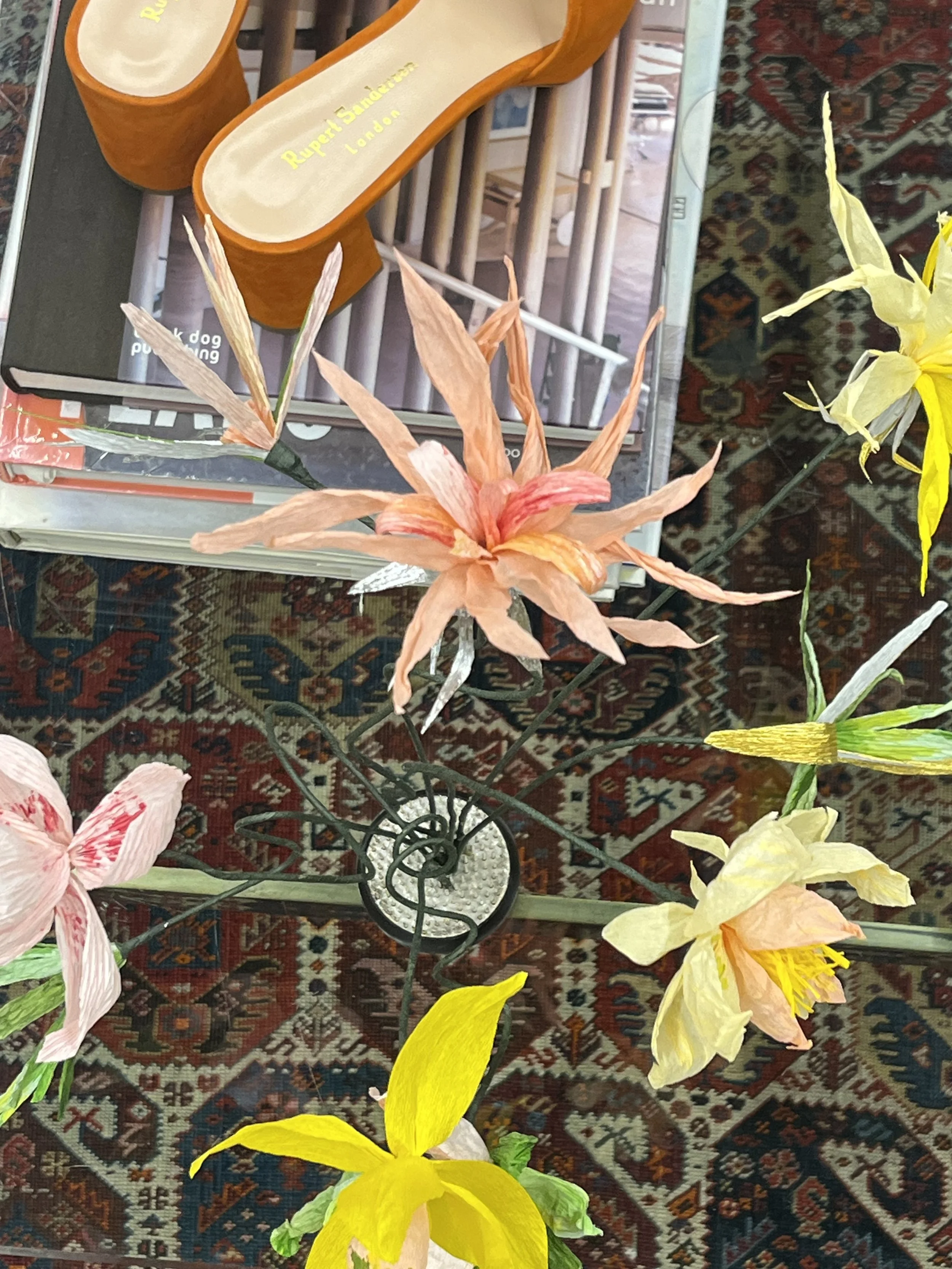 A display of decorative artificial flowers, including pink, yellow, and orange lilies by Emmeli Kimhi, arranged on a table with a patterned red and blue rug underneath. Part of a small decorative metal bicycle stand is visible.