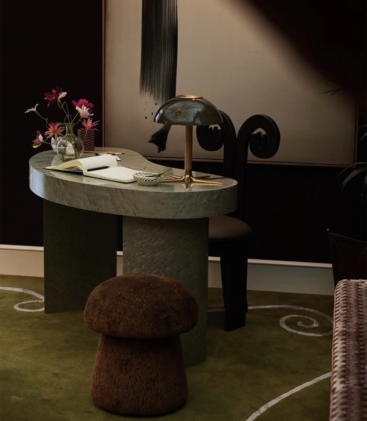 A round, greenish marble table with a vase of pink paper flowers by Emmeli Kimhi, small bowl, and a black and gold desk lamp. Behind the table is a black chair with a scroll design backrest, and part of a dark art piece is visible on the wall. 