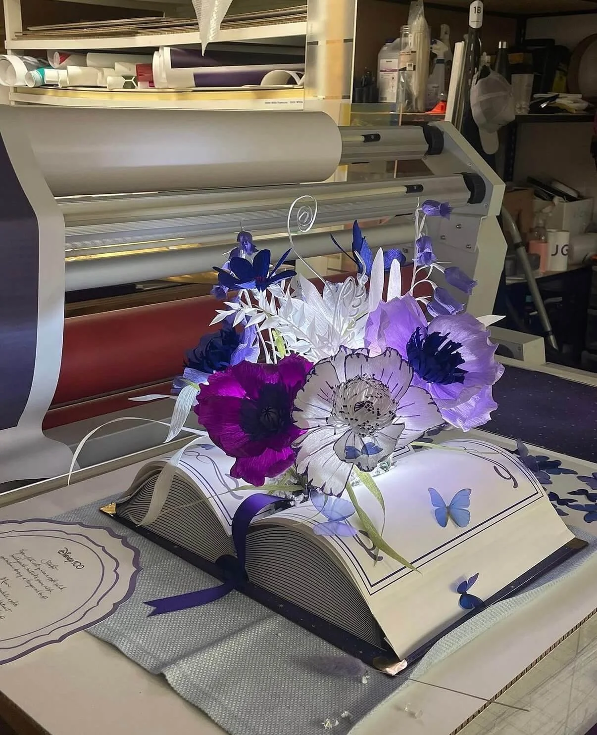 A floral arrangement by Emmeli Kimhi with paper flowers and butterflies on an open book, placed on a table with a gray cloth, in front of a vinyl cutting machine in a workshop.