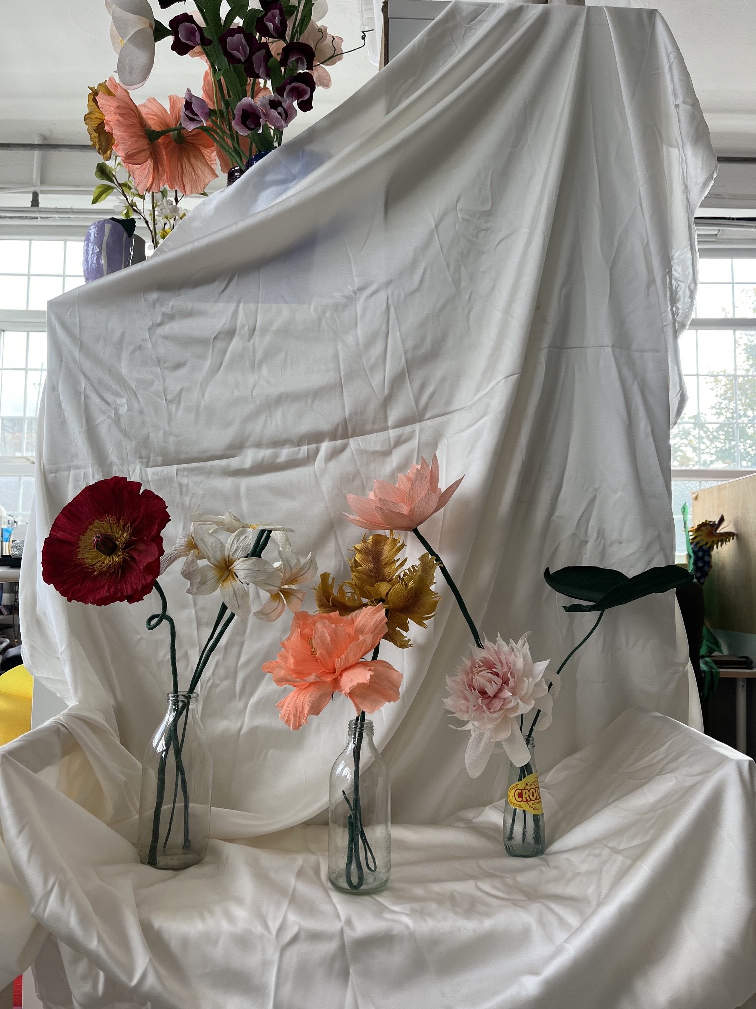 Flowers by Emmeli Kimhi in vases on a white draped surface, with a backdrop of a window and an indoor setting.
