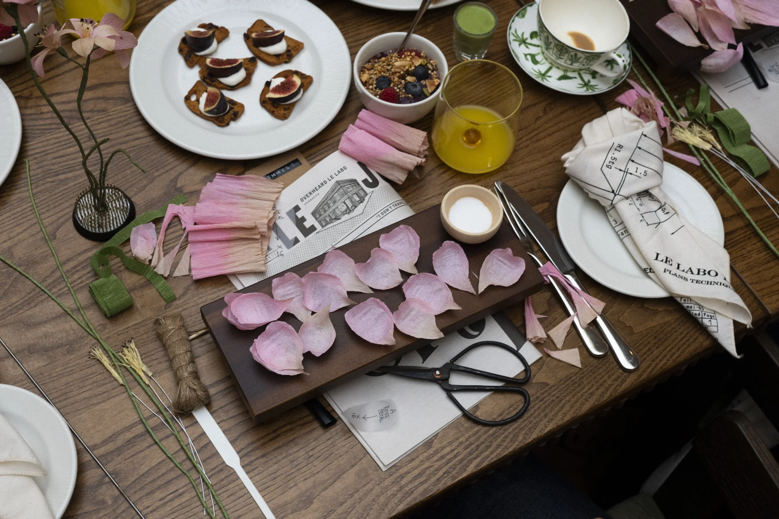 Emmeli Kimhi workshop. A woodworking workspace with pink flower petals, scissors, and pink and green floral wire on a wooden table, along with a few plates of snacks, a small bowl of sugar, and a glass of yellow drink.