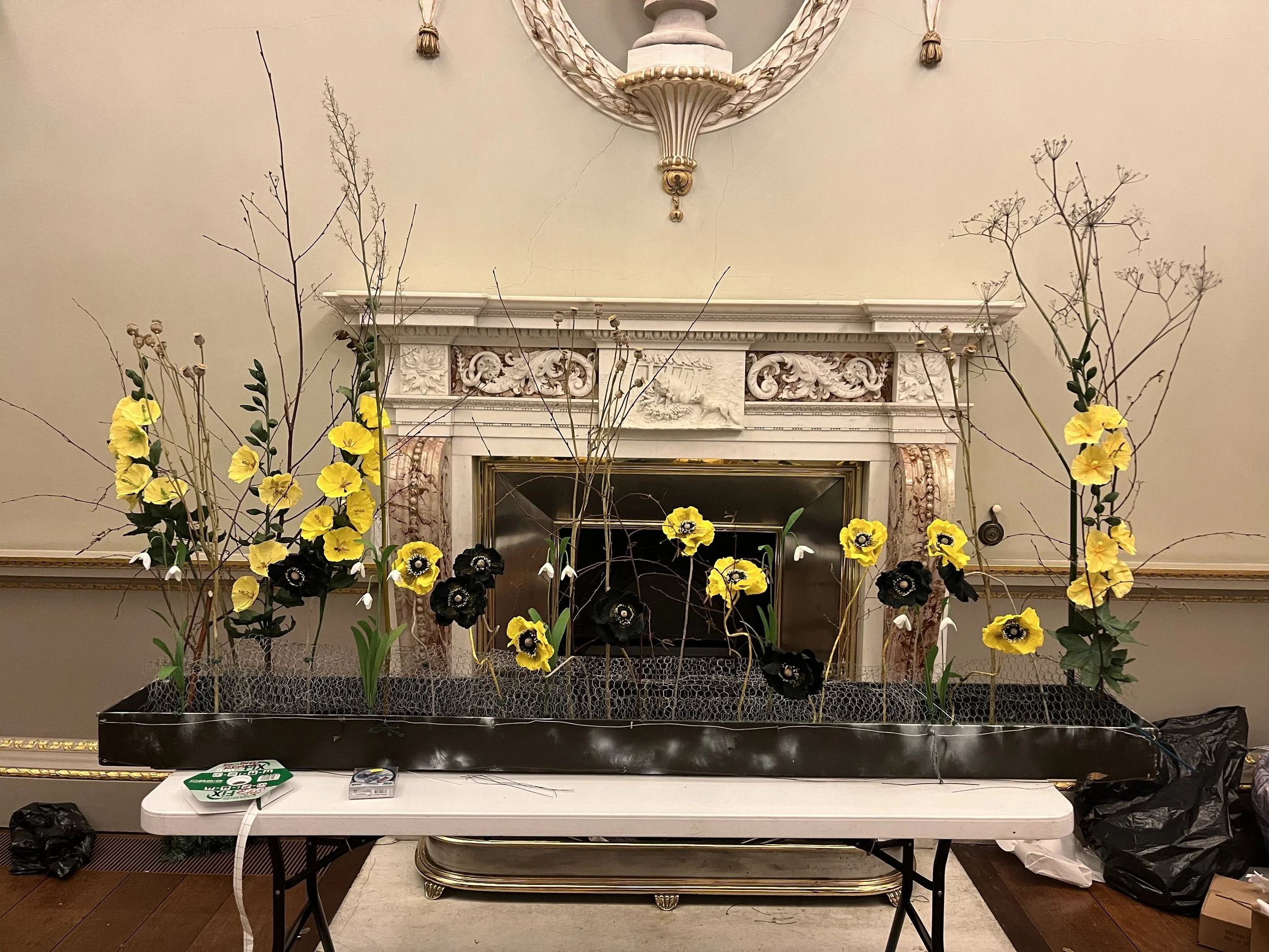 The image shows decorative arrangement of yellow and black flowers with green leaves and budding branches in a black container placed on a white table in front of a vintage ornate marble fireplace and a large round mirror with tassel ornaments hangin