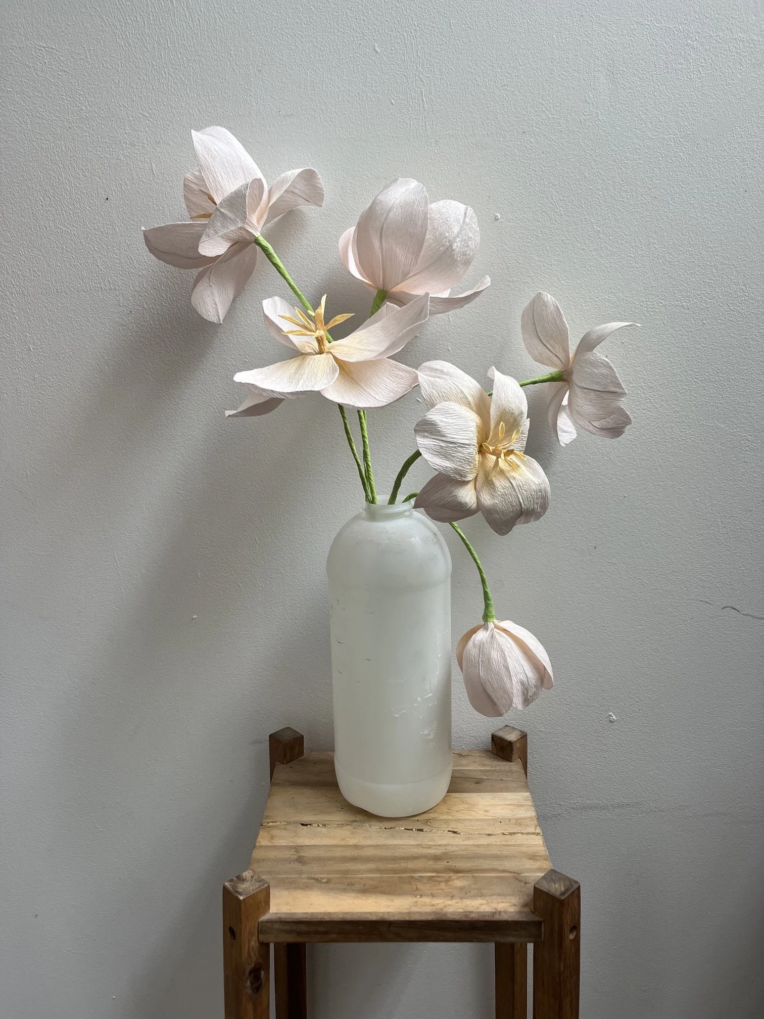 A white vase with blush pink and cream-colored paper flowers by Emmeli Kimhi, placed on a rustic wooden table against a plain gray wall.