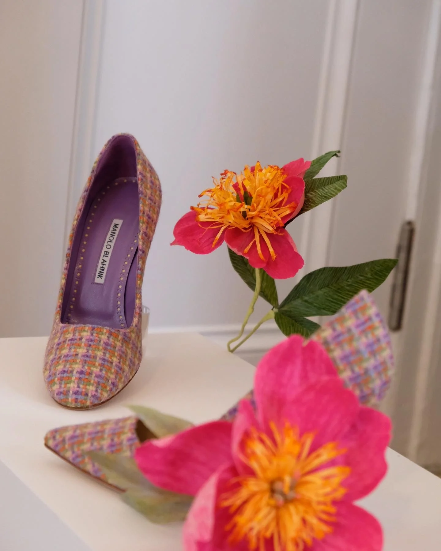 A colorful plaid high-heeled shoe by Manolo Blahnik, placed on a white surface beside pink and orange paper flowers by Emmeli Kimhi