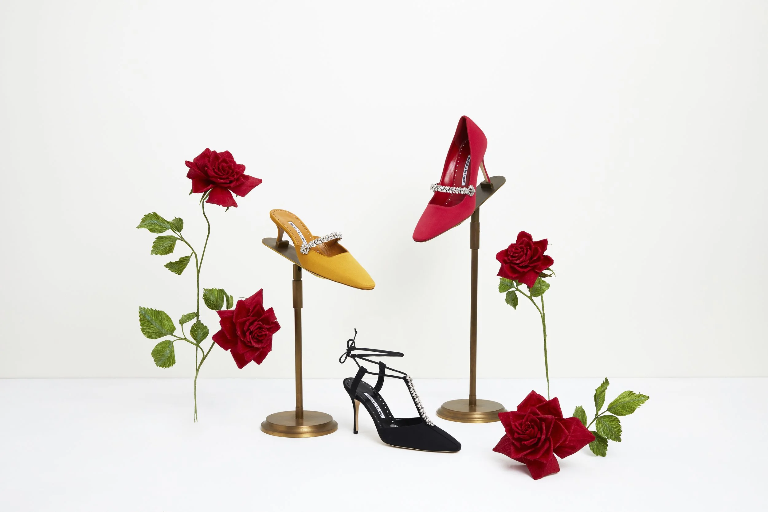 Display of four colorful high-heeled shoes on stands, surrounded by red paper roses  by Emmeli Kimhi with green leaves, set against a plain white background.