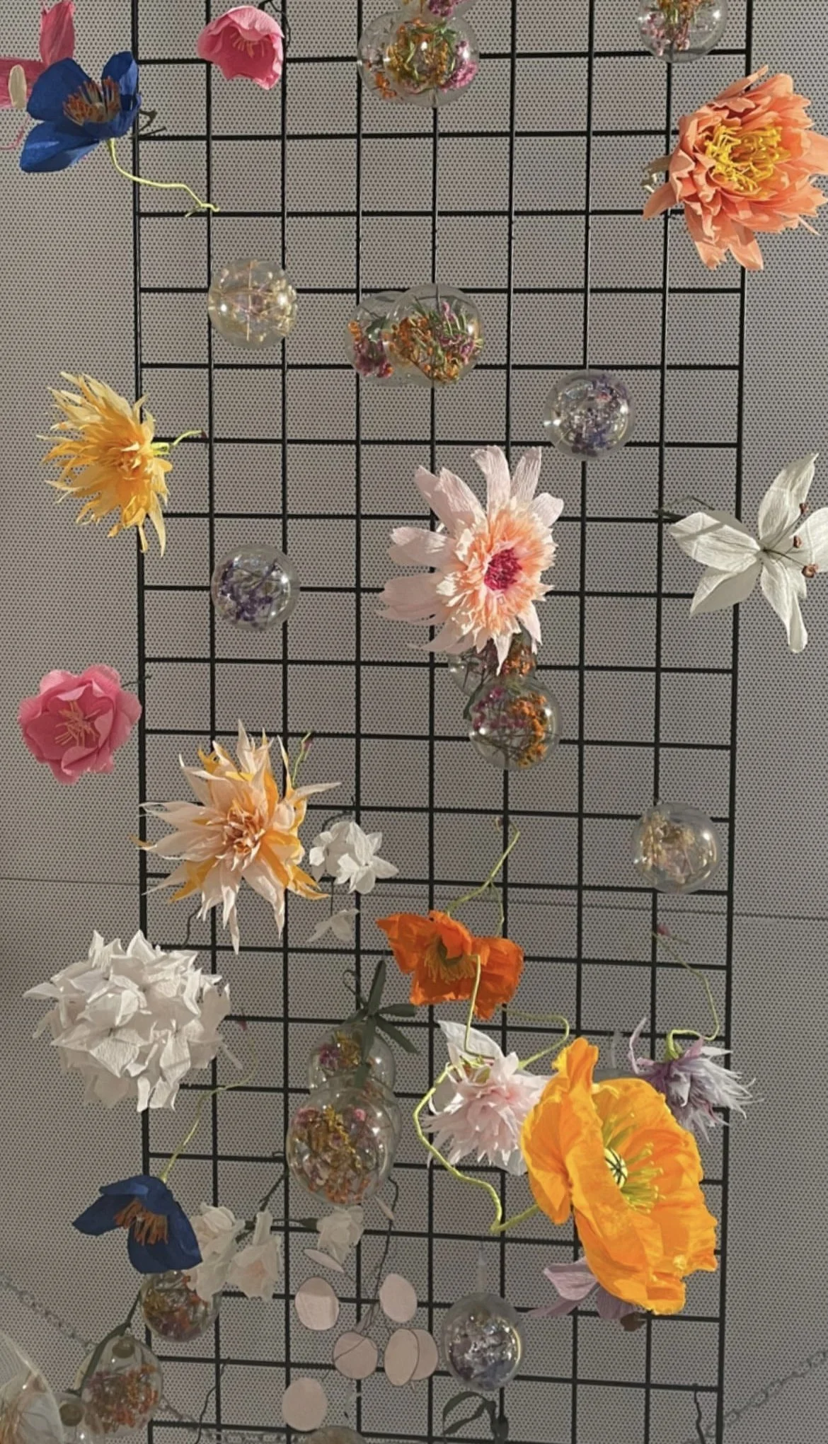 Decorative display of colorful artificial flowers by Emmeli Kimhi and glass orbs attached to a black wire grid.