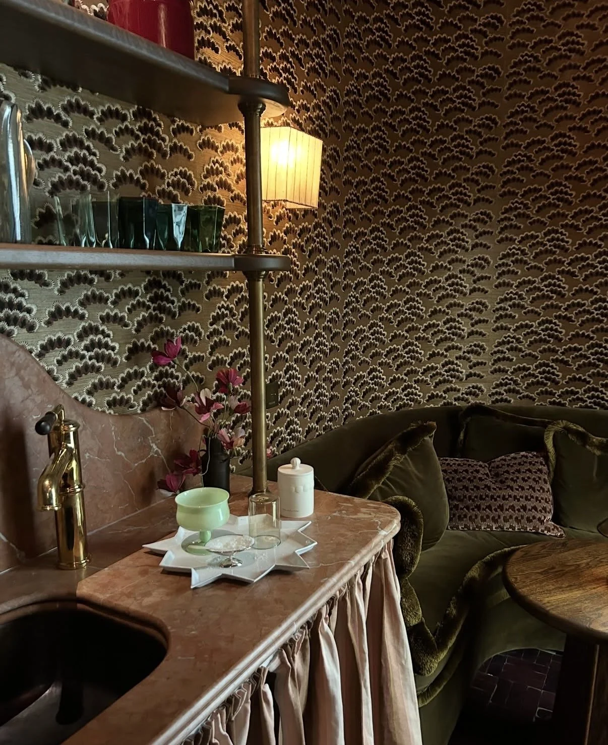 Interior of a vintage-style living room or bar area with patterned wallpaper, a marble countertop with a flower arrangement by Emmeli Kimhi, glassware, and a cream-colored container. 