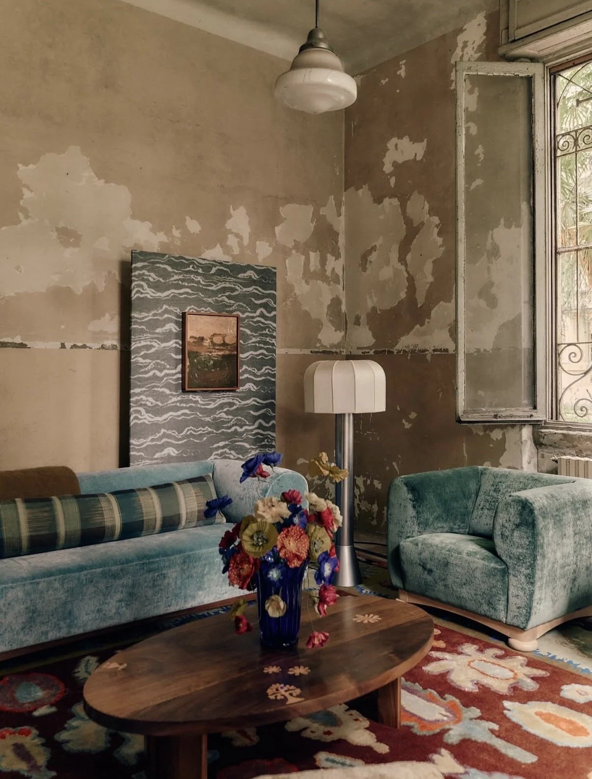 Emmeli Kimhi’s vibrant hand-painted paper poppy bouquet in a blue glass vase, displayed on a wooden oval coffee table in Studio Ashby’s installation at Alcova Milan. The colourful arrangement sits in a room with distressed walls, teal velvet seating,