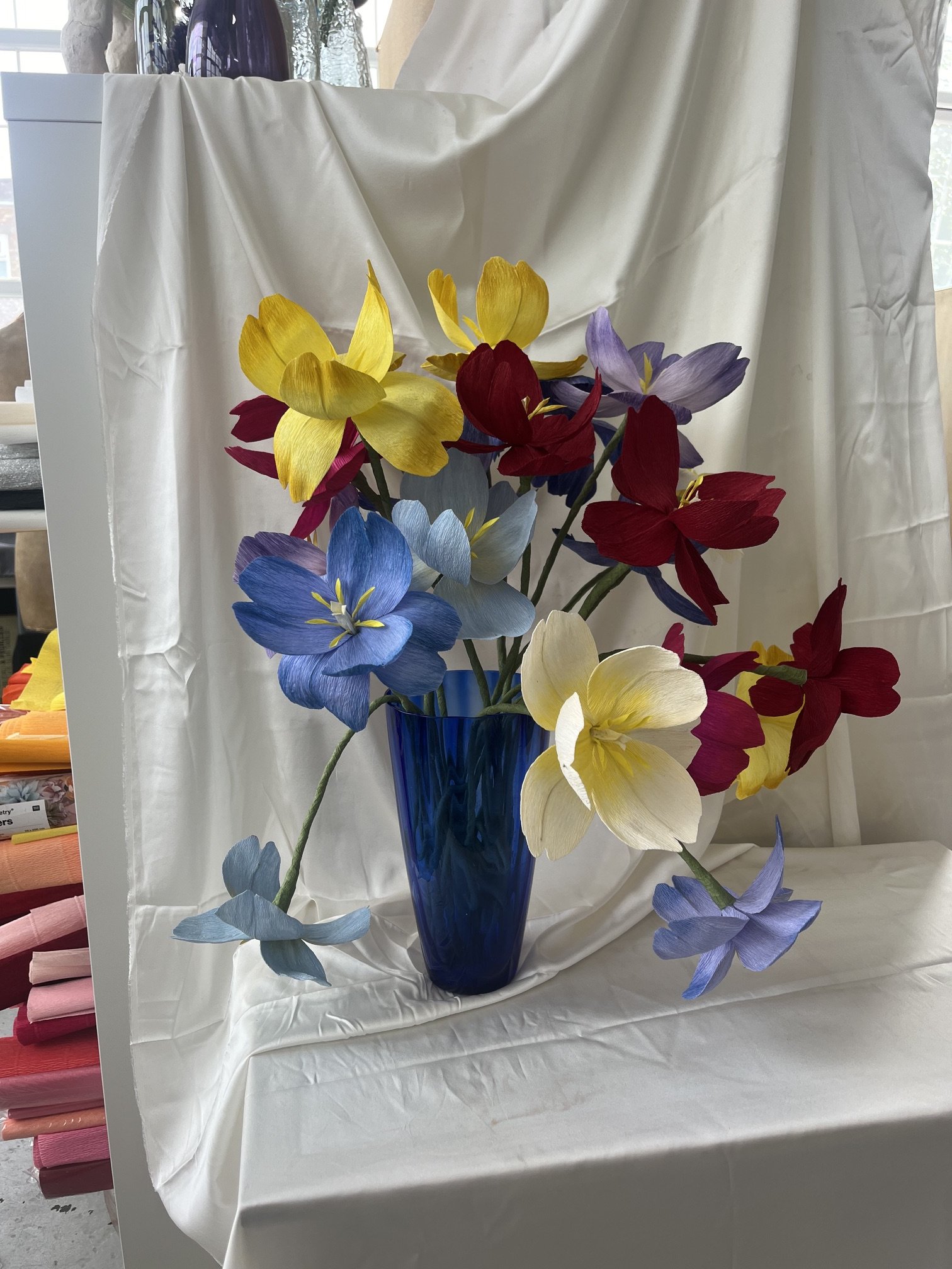A bouquet of colorful paper flowers by Emmeli Kimhi in a blue glass vase on a table with a white tablecloth. The flowers are yellow, red, purple, blue, and cream, with some petals falling around the vase.