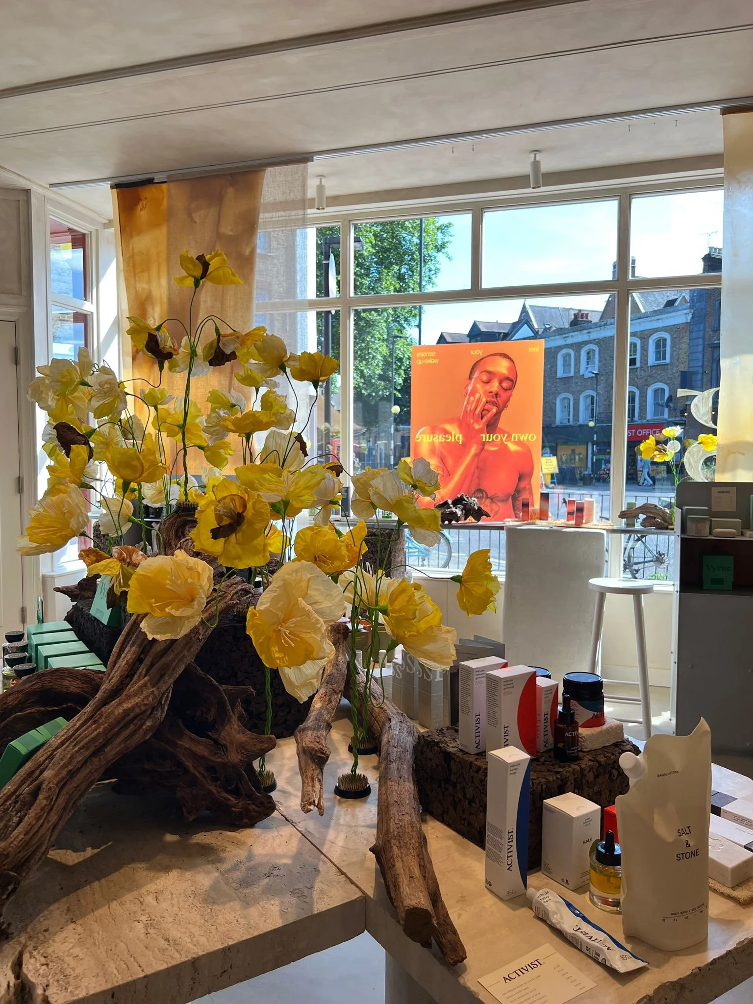 Interior of a storefront with a large window, display of yellow flowers by Emmeli kimhi and natural wood decorations, skincare products on the counter, and a bright street scene outside. A promotional poster with a woman yawning
