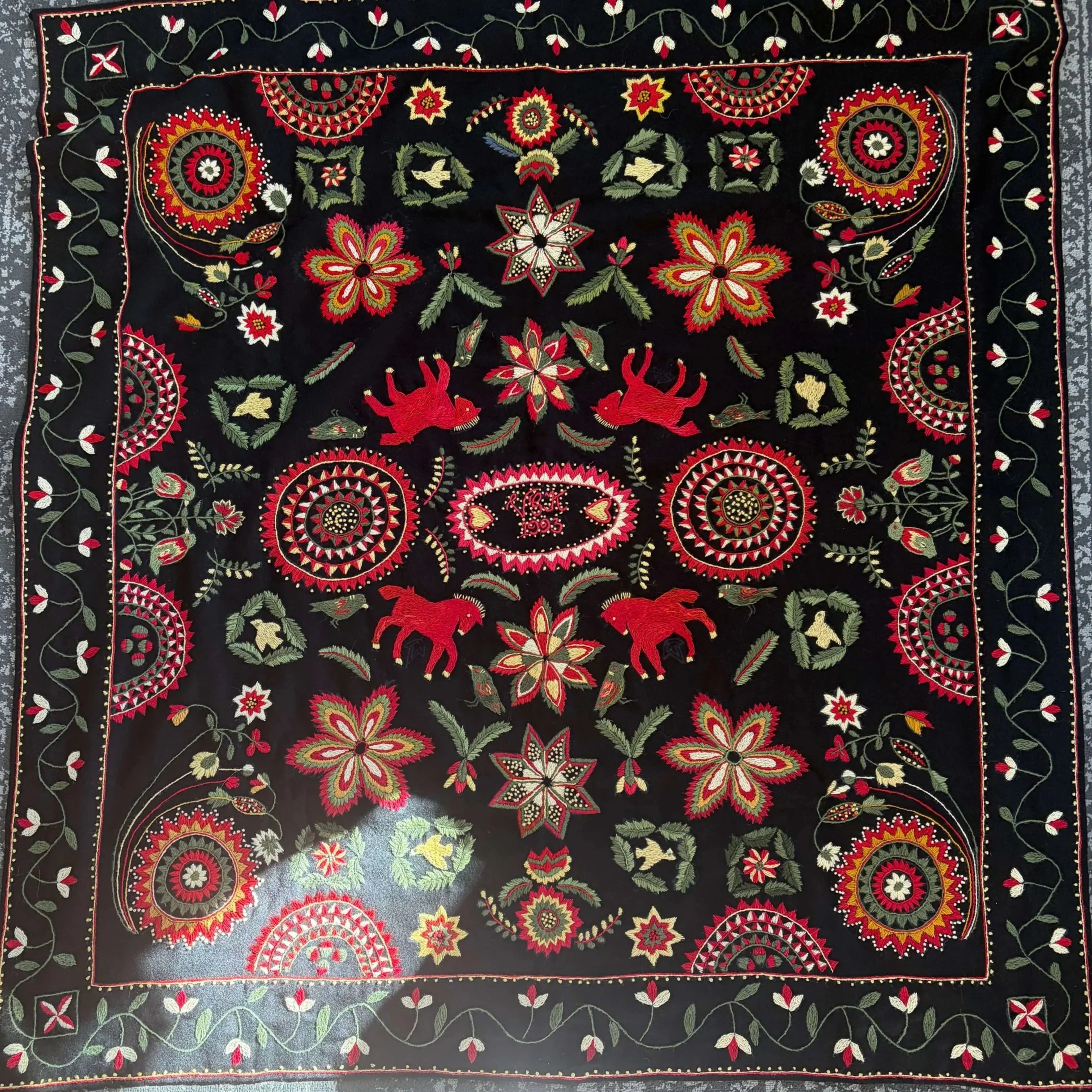 A full skånsk yllebroderi wool embroidery in black, red, cream, and olive green, with repeating floral medallions, horses, and birds, and an initialled oval at the centre.