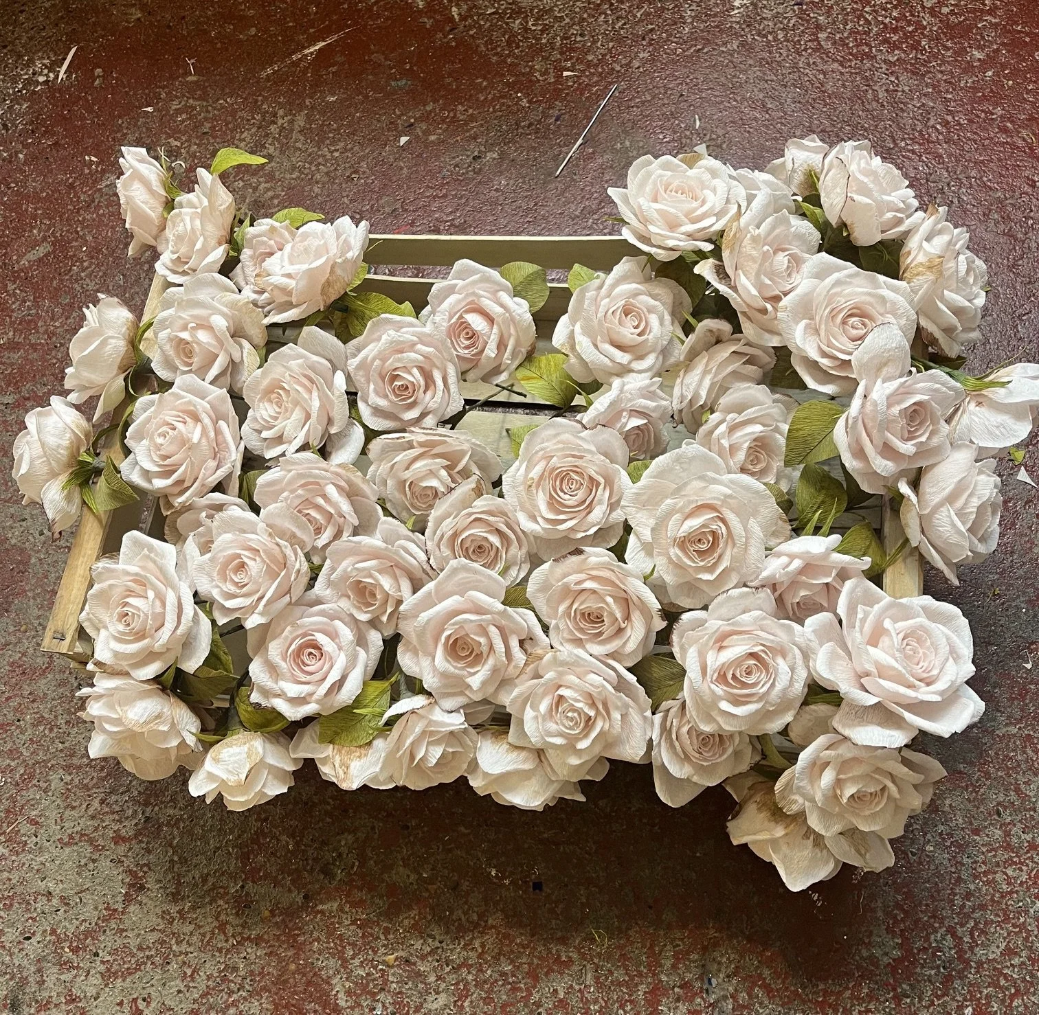 A crate filled with pink paper roses by Emmeli Kimhi laying on a red textured surface.