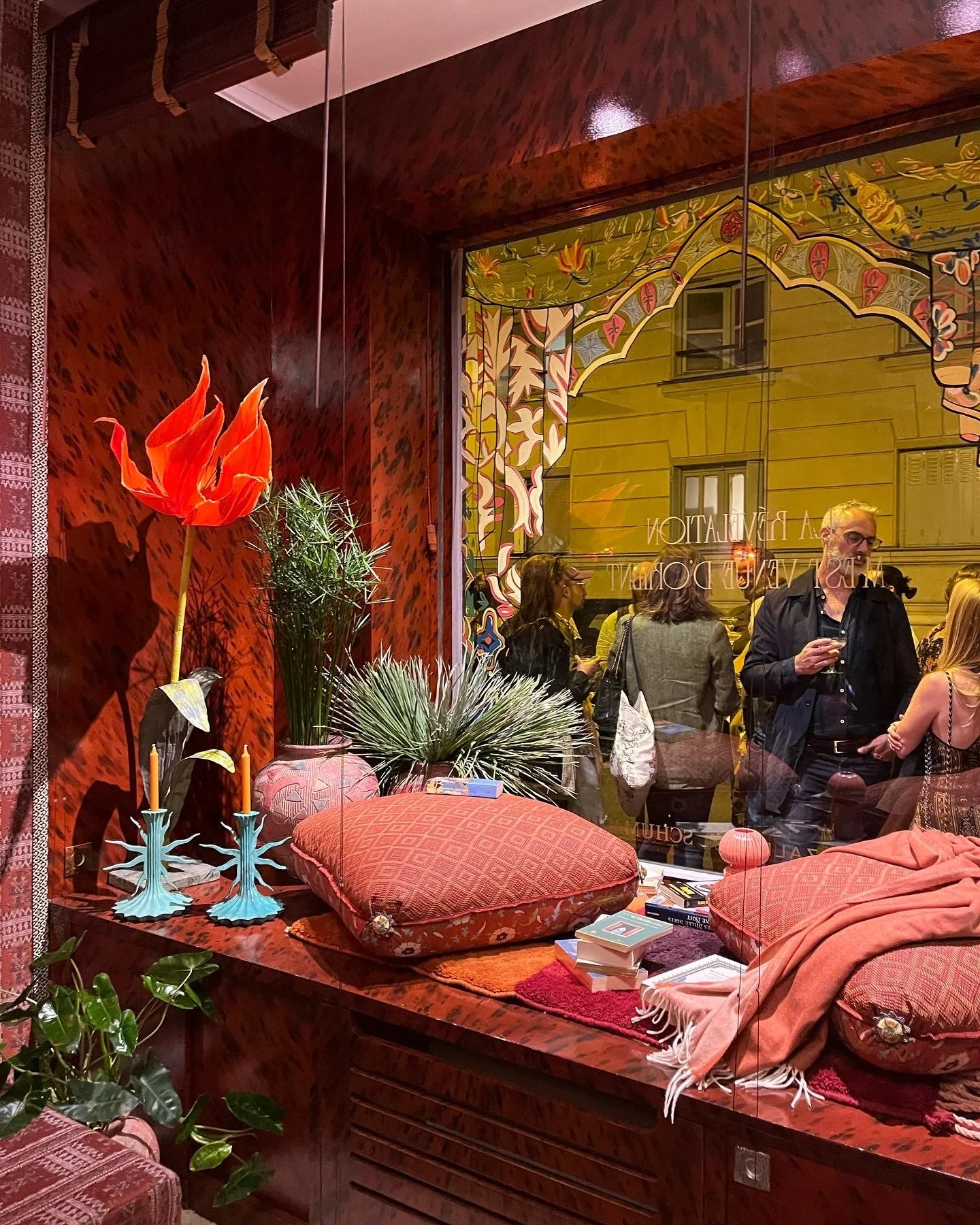 A cozy room with red patterned walls and sofa cushions, decorated with a plant, flower by Emmeli Kimhi, books, and candles, seen through a window with reflections of people socializing outside.