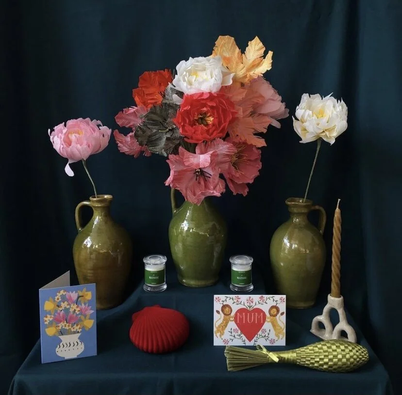 A memorial display with three green vases holding paper flowers by Emmeli, two candles, a red knitted item, a Mother's Day card, a decorative yellow-green object, and a white ceramic candle holder with a twisted candle, set against a dark background.