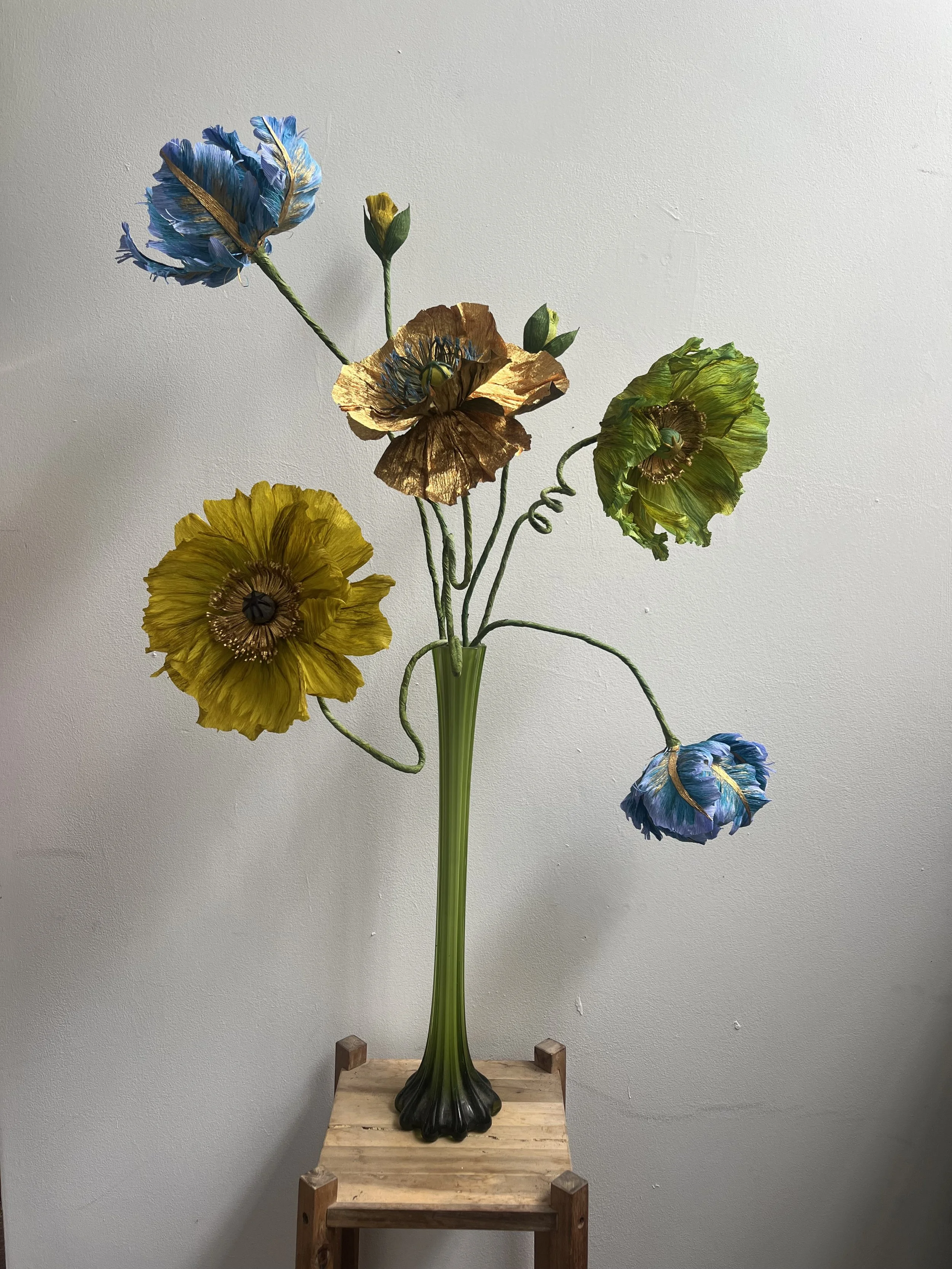 A tall glass bouquet with five colorful artificial flowers by Emmeli Kimhi, including blue, yellow, and green, arranged on a small wooden stand against a plain white wall.