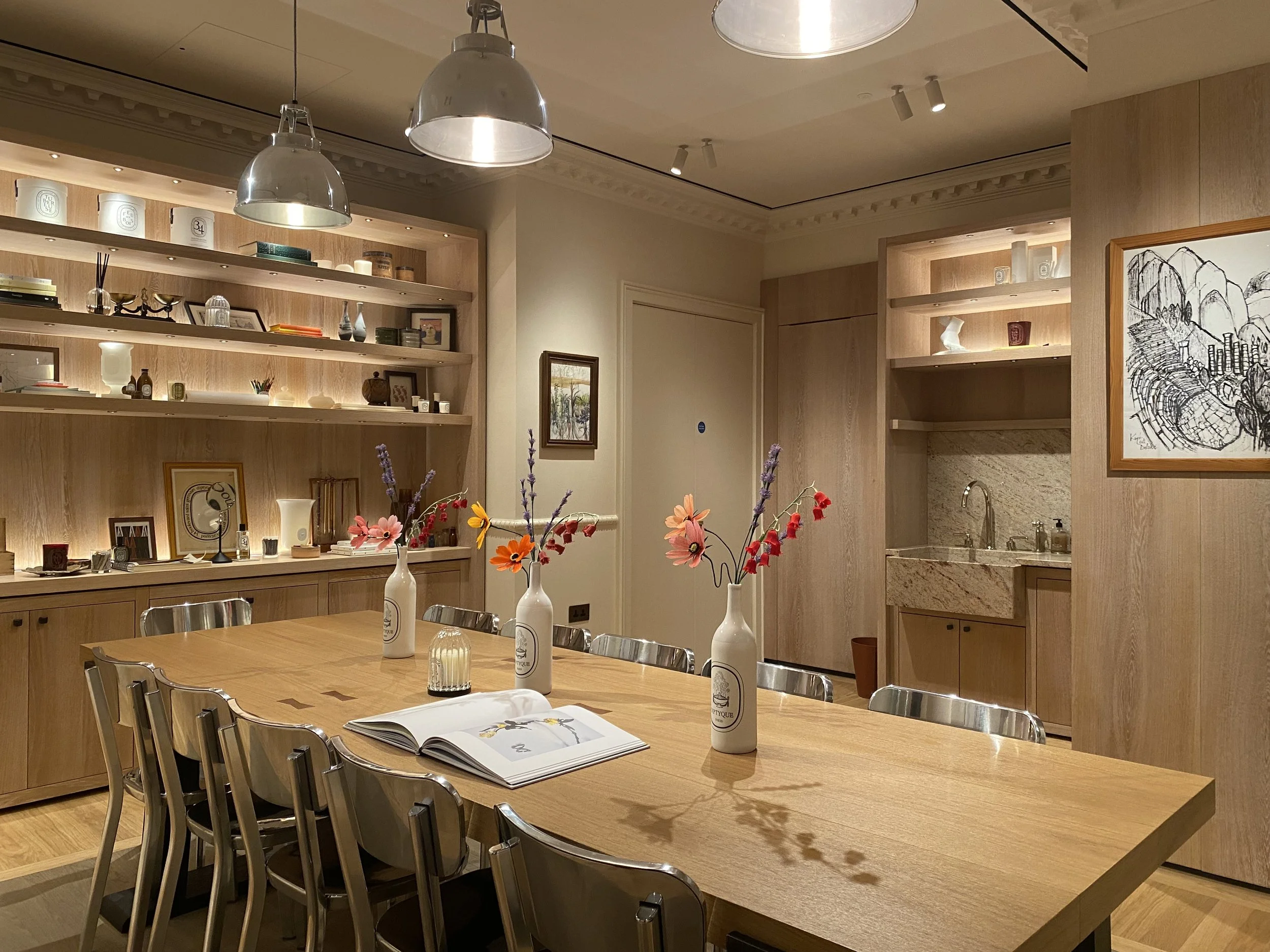 A wooden dining table with metal chairs, decorated with white vases and colourful paper flowers by Emmeli Kimhi for Diptyque, and a book open on it, in a well-lit room with wood shelves and artwork on the walls.