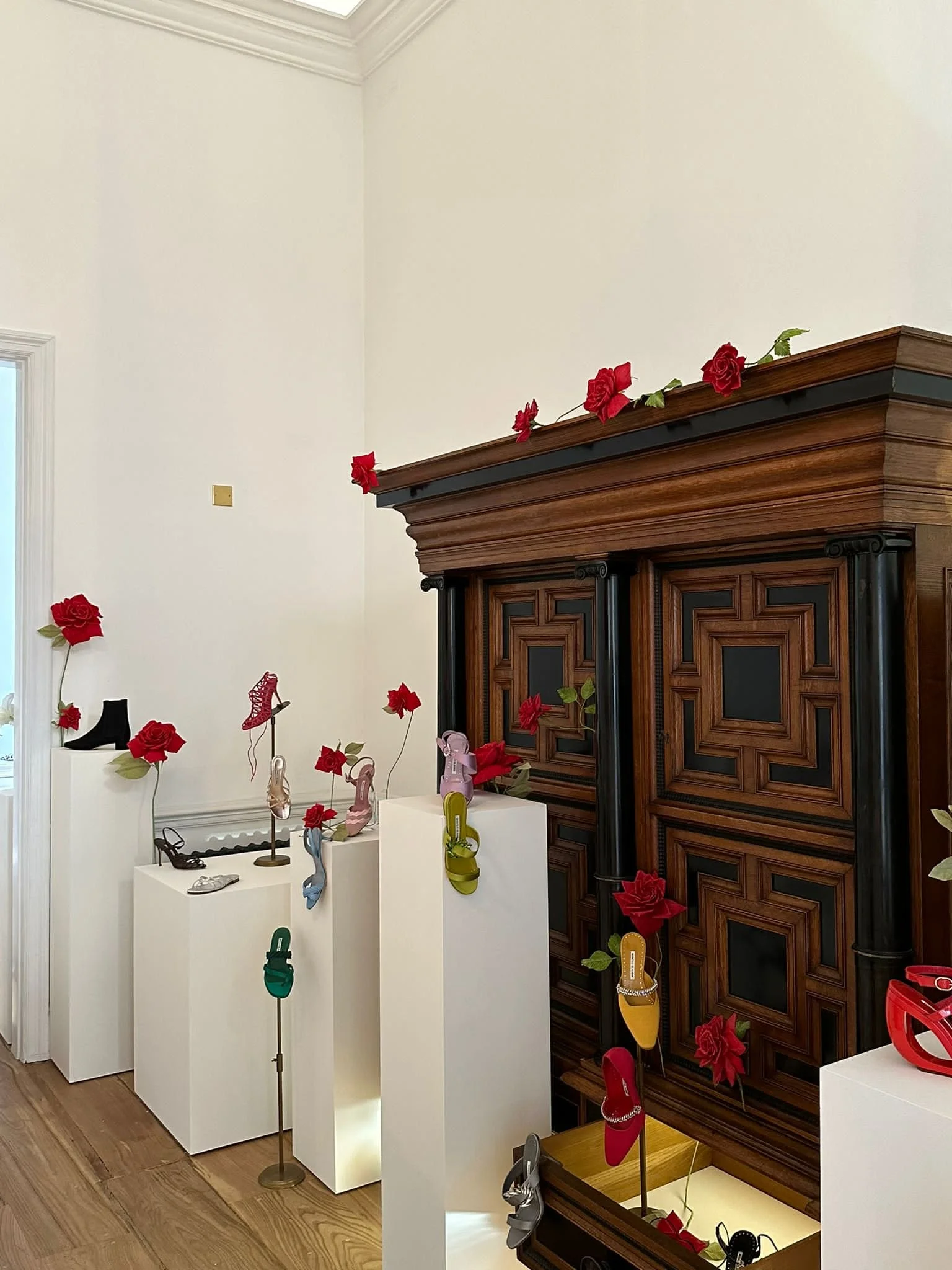 Display of various colorful women's shoes and accessories arranged on white and wooden pedestals in a room with white walls and wooden floor.