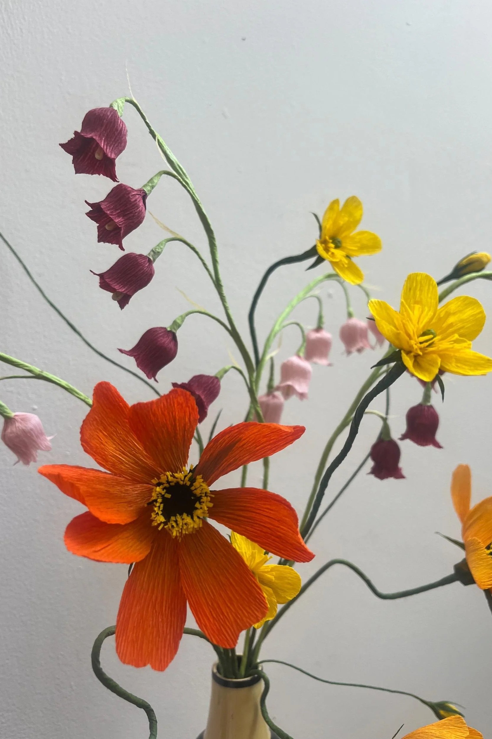 Artistic handmade paper flower arrangement with bold orange cosmos, bright yellow blooms and delicate hanging bells in a ceramic vase by Emmeli Kimhi