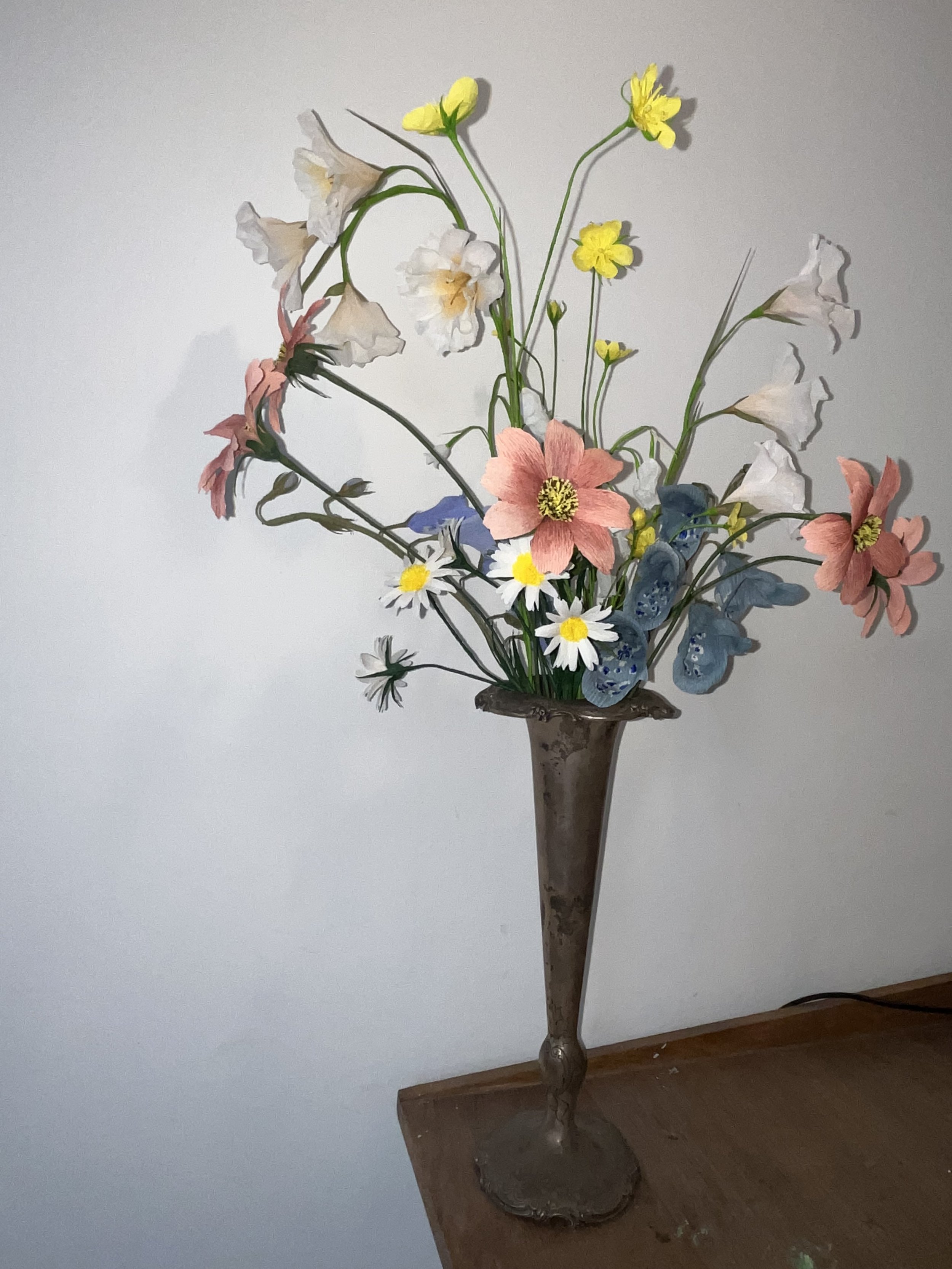 A paper flower arrangement by Emmeli Kimhi in a decorative metal vase with a wooden base, featuring pink, white, yellow, and purple flowers, and green foliage, placed on a wooden surface against a plain light-colored background.