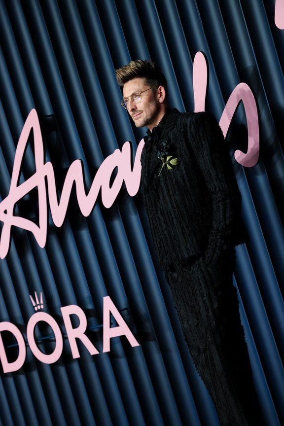 Henry Holland with glasses and styled hair standing in front of a dark blue ridged backdrop with pink text that reads 'Shangri-La' and 'Dora'