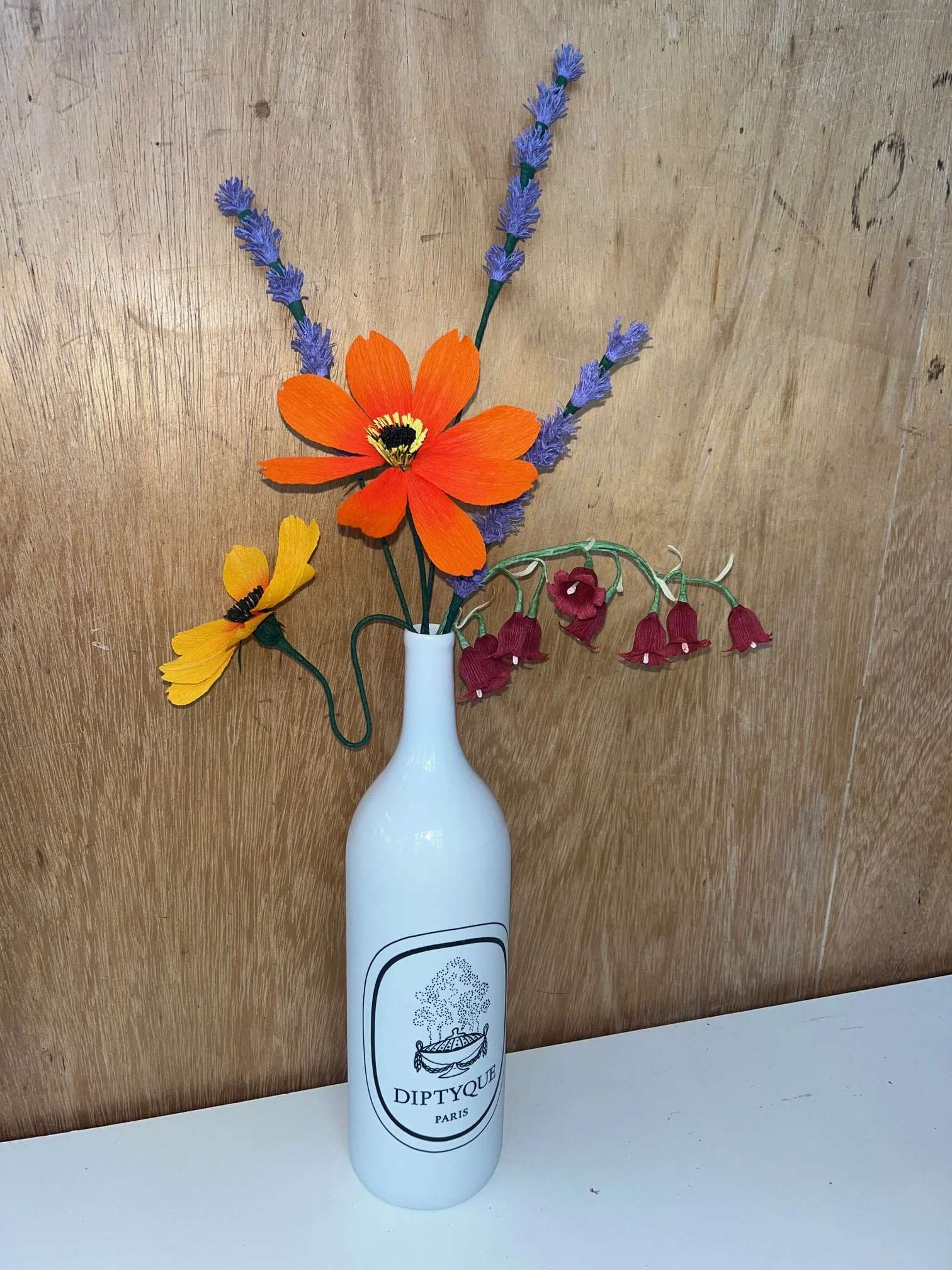 A white bottle with a Diptyque Paris logo in front of a wooden background, holding an arrangement of artificial flowers by Emmeli Kimhi including large orange and smaller yellow flowers, with purple lavender-like stems and red bell-shaped flowers.