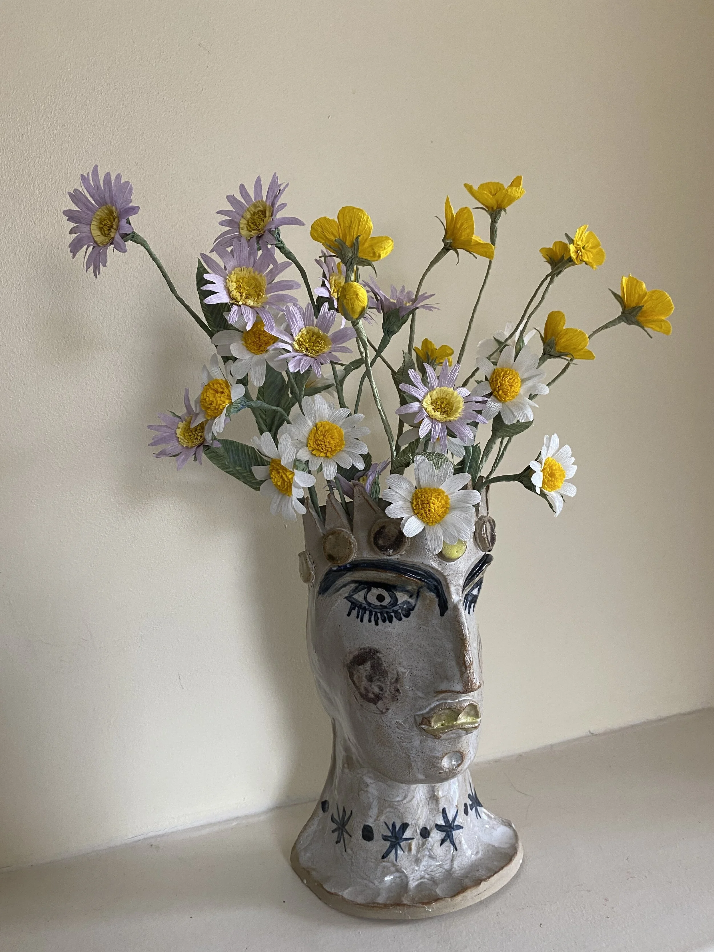 A ceramic vase in the shape of a woman's face with painted features, holding a bouquet of purple, white, and yellow paper flowers by Emmeli Kimhi, placed against a plain beige wall.